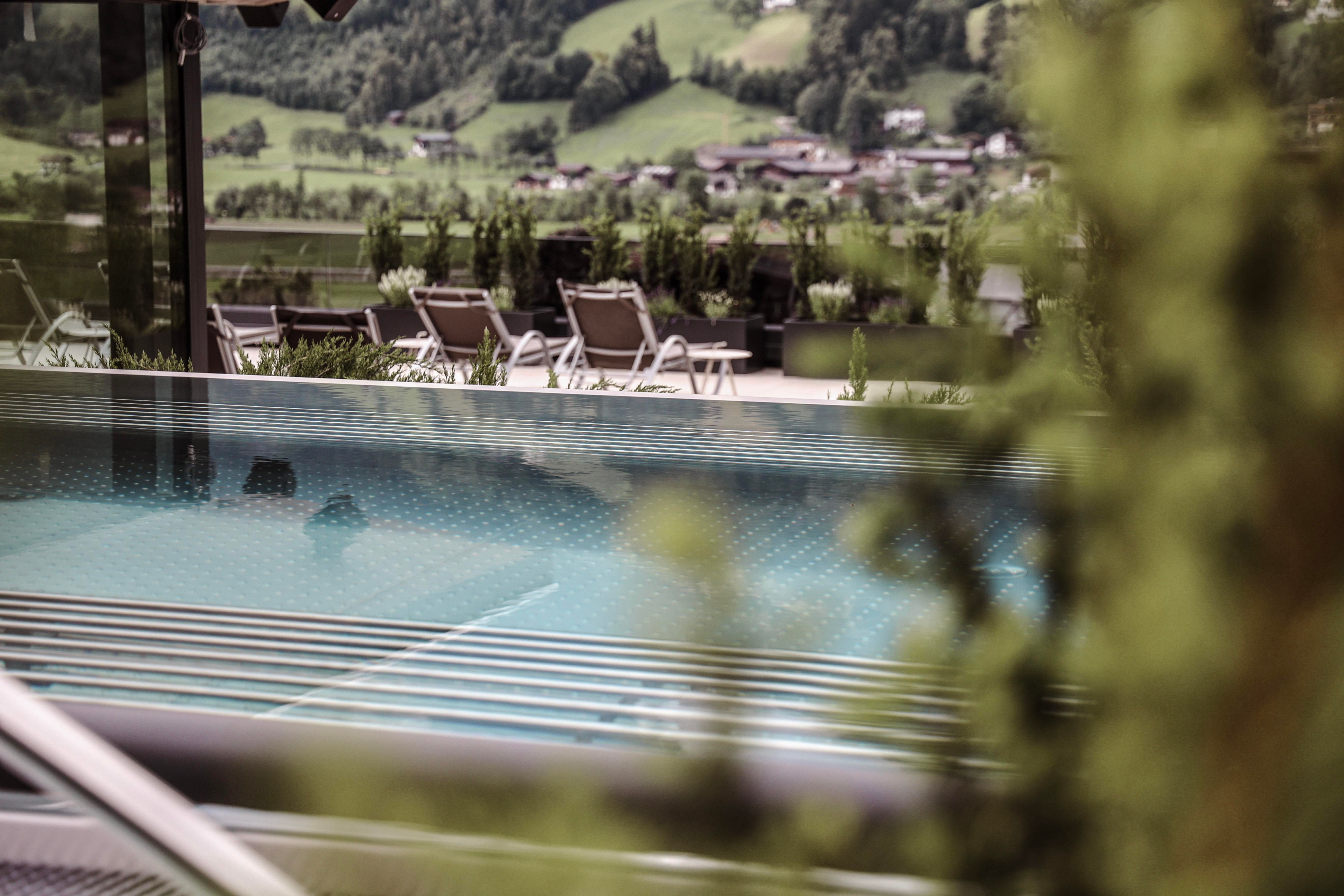 A modern pool with a beautiful view of the surrounding hills. Sun loungers are set up in the background, surrounded by plants.