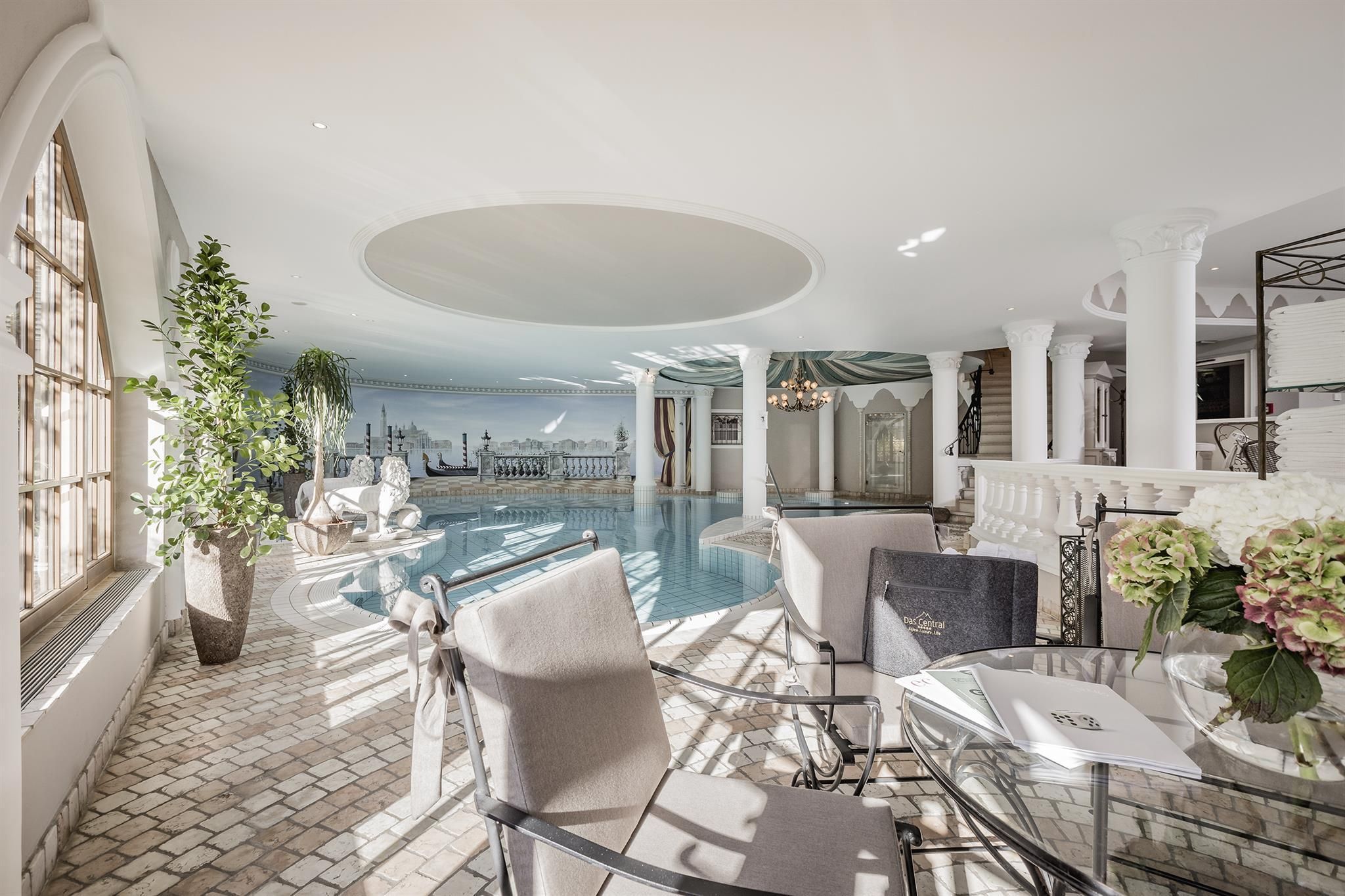 An elegant indoor pool with large windows and plenty of daylight. There are comfortable seating areas and decorative plants around the pool area.