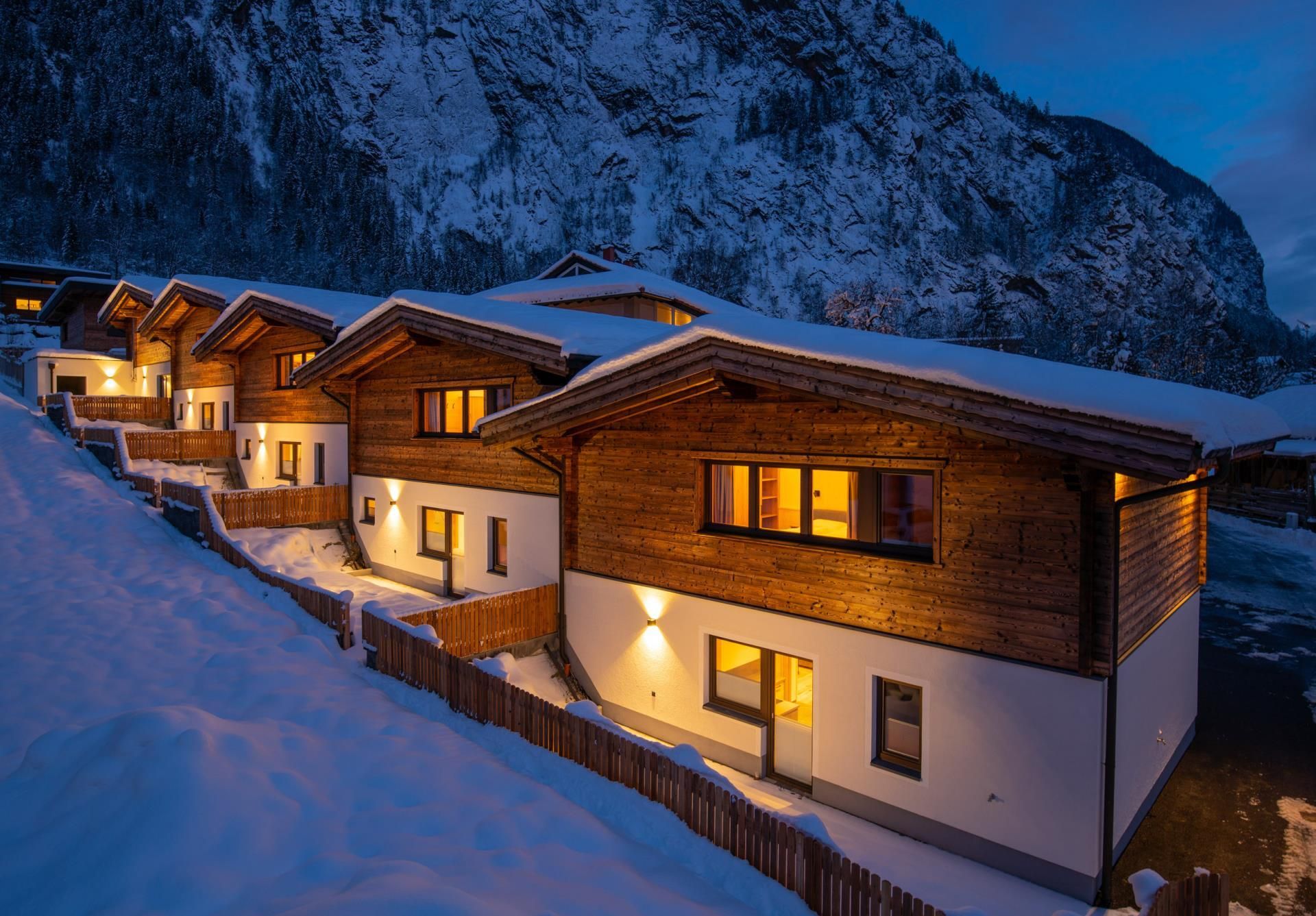 A group of modern wooden chalets in the snow, surrounded by mountains. The chalets are illuminated at night, creating a cozy atmosphere.