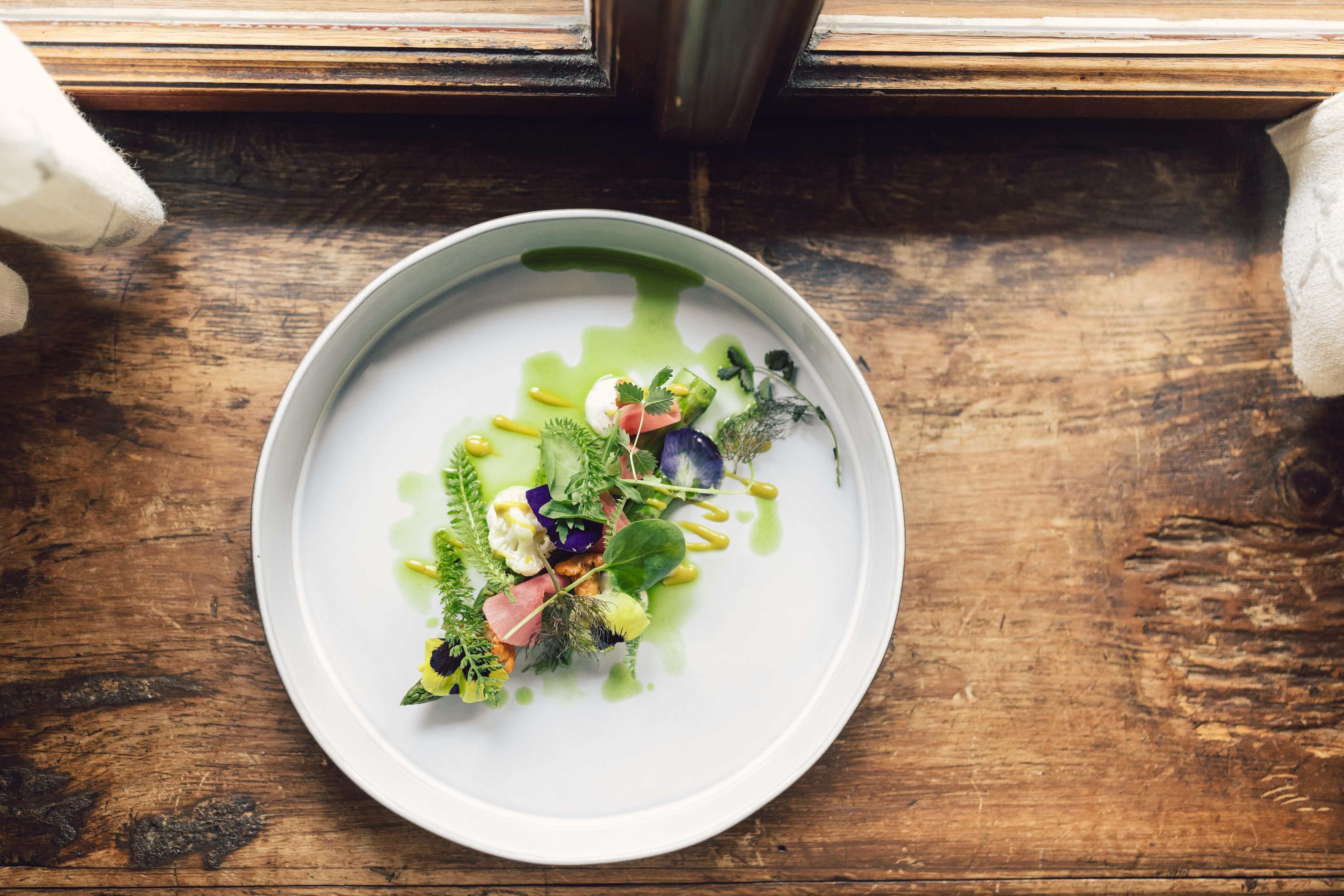 An artfully arranged plate with fresh ingredients, including vegetables and herbs. The plate is on a rustic wooden table next to a window.