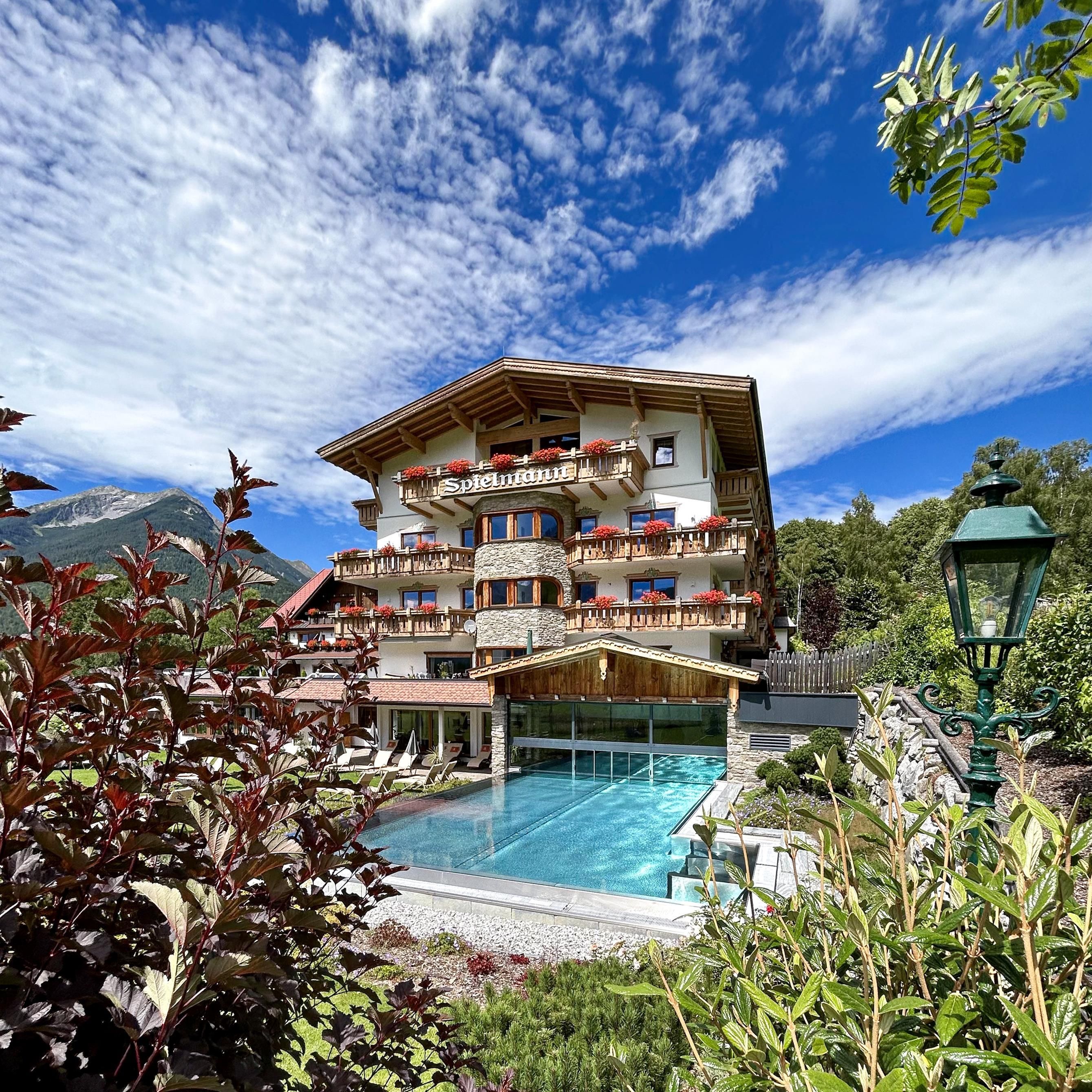 A beautiful hotel with balconies and a pool in the foreground. The sky is clear with some clouds and the surroundings are green and inviting.