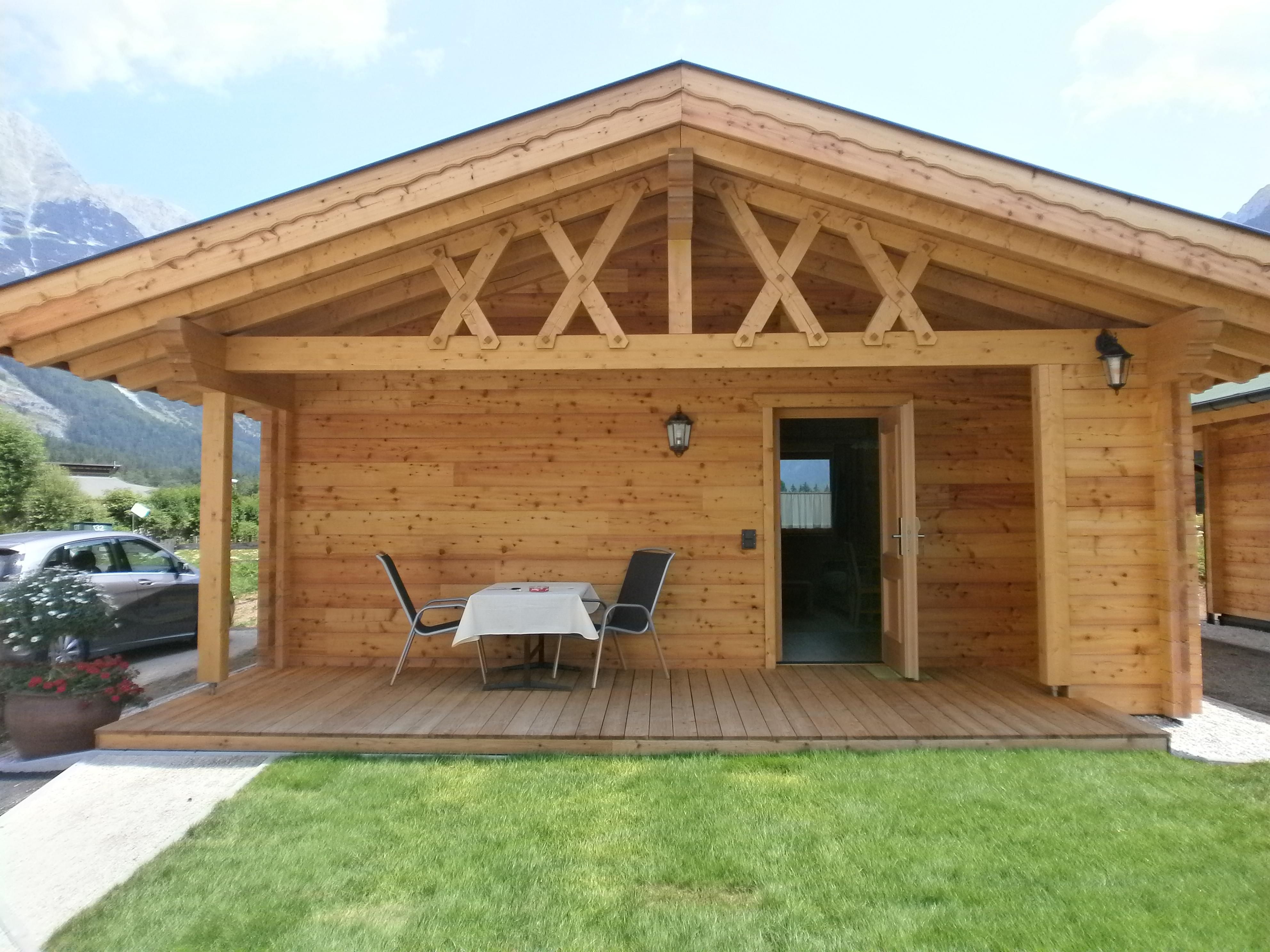 A cozy wooden house with a covered entrance area. In front of the door, there is a table with chairs on a green lawn.