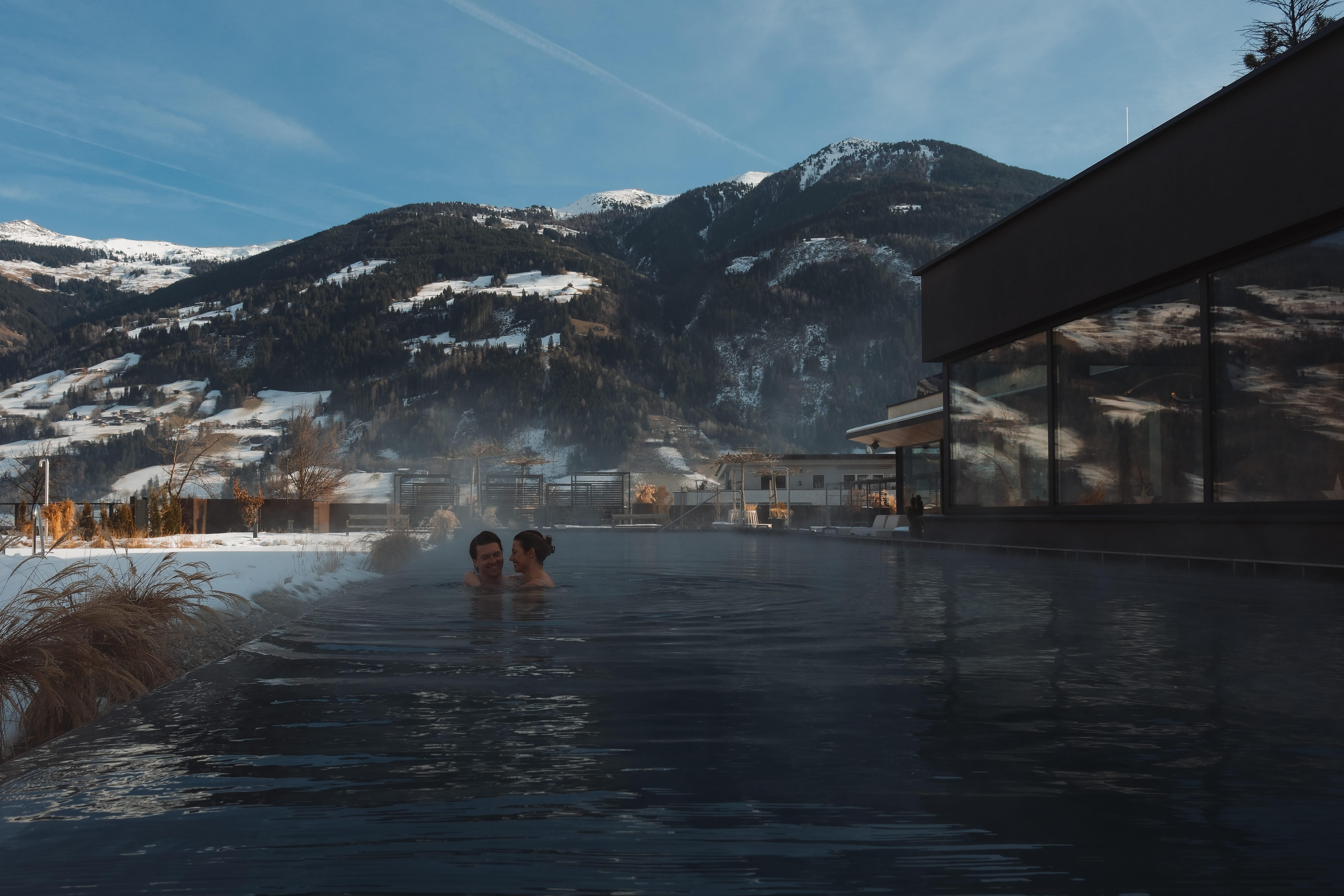 A couple relaxes in a heated pool overlooking snow-capped mountains. The sky is clear, and the atmosphere is calm and refreshing.