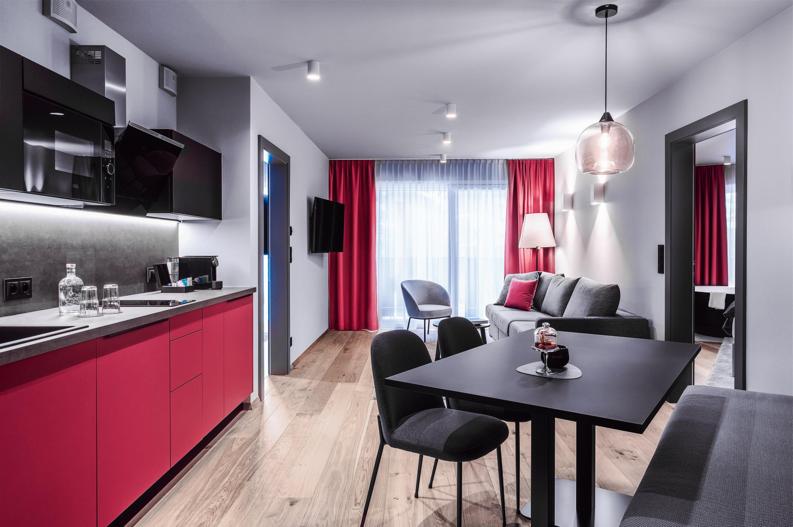 A modern apartment with an open kitchen and a dining area. The decor is in black and red, and there is plenty of daylight.