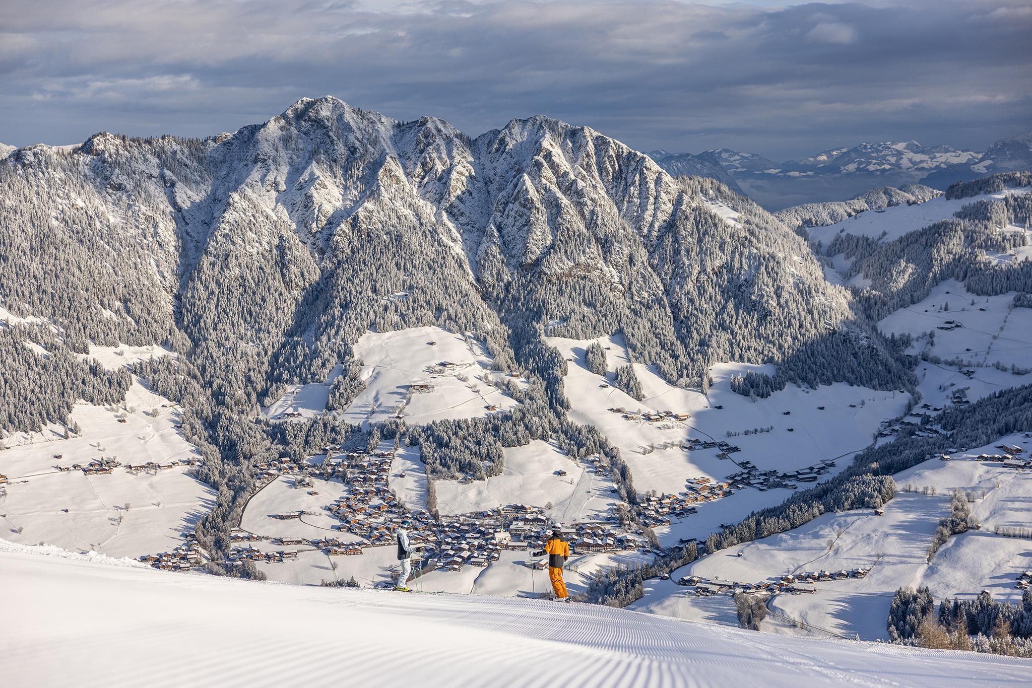 A snowboarder stands on a snow-covered mountain peak with impressive, snow-covered mountains in the background. The landscape is wintery and idyllic.
