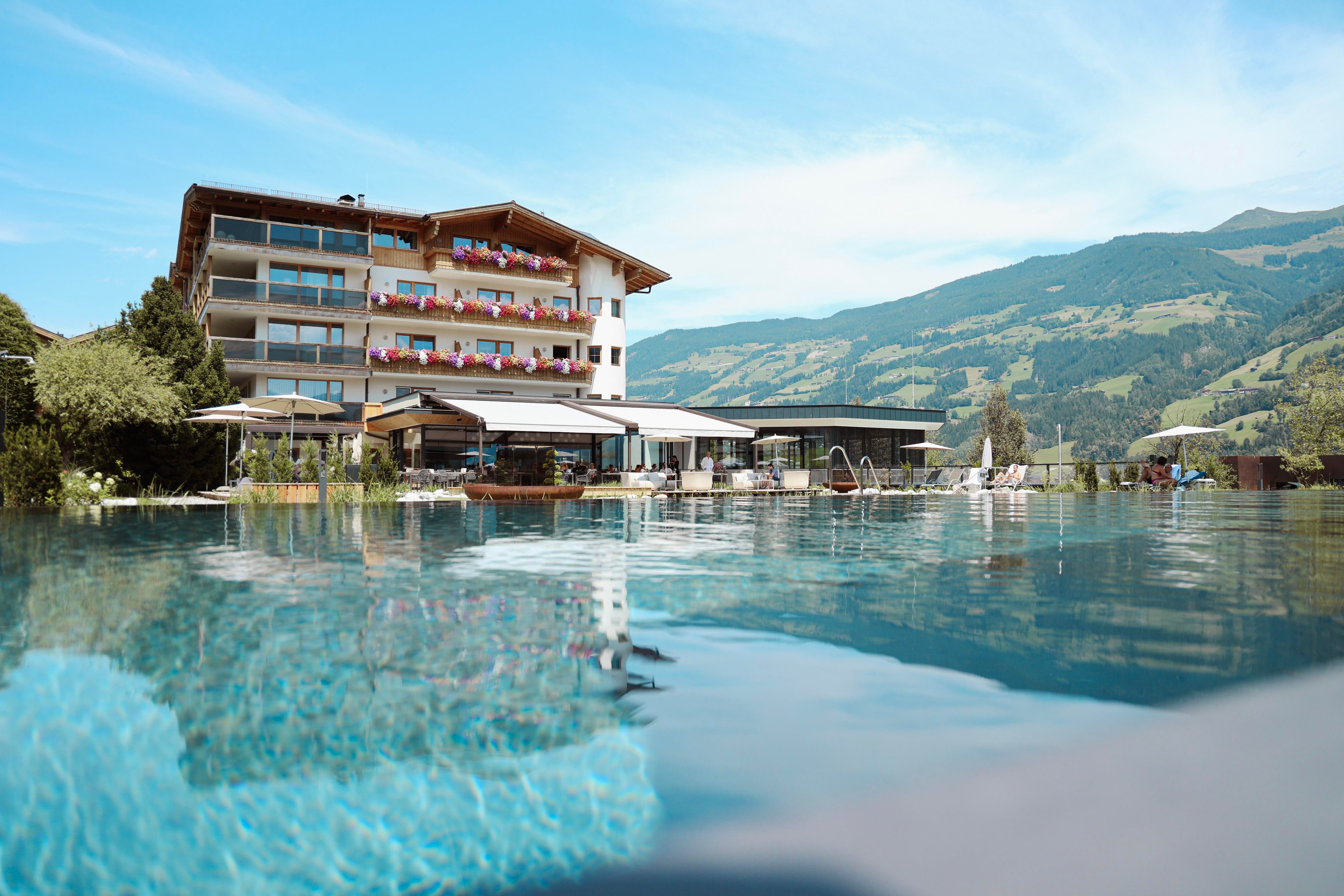 A modern hotel with a large pool nestled among green mountains. The terrace is comfortably furnished and invites you to relax.