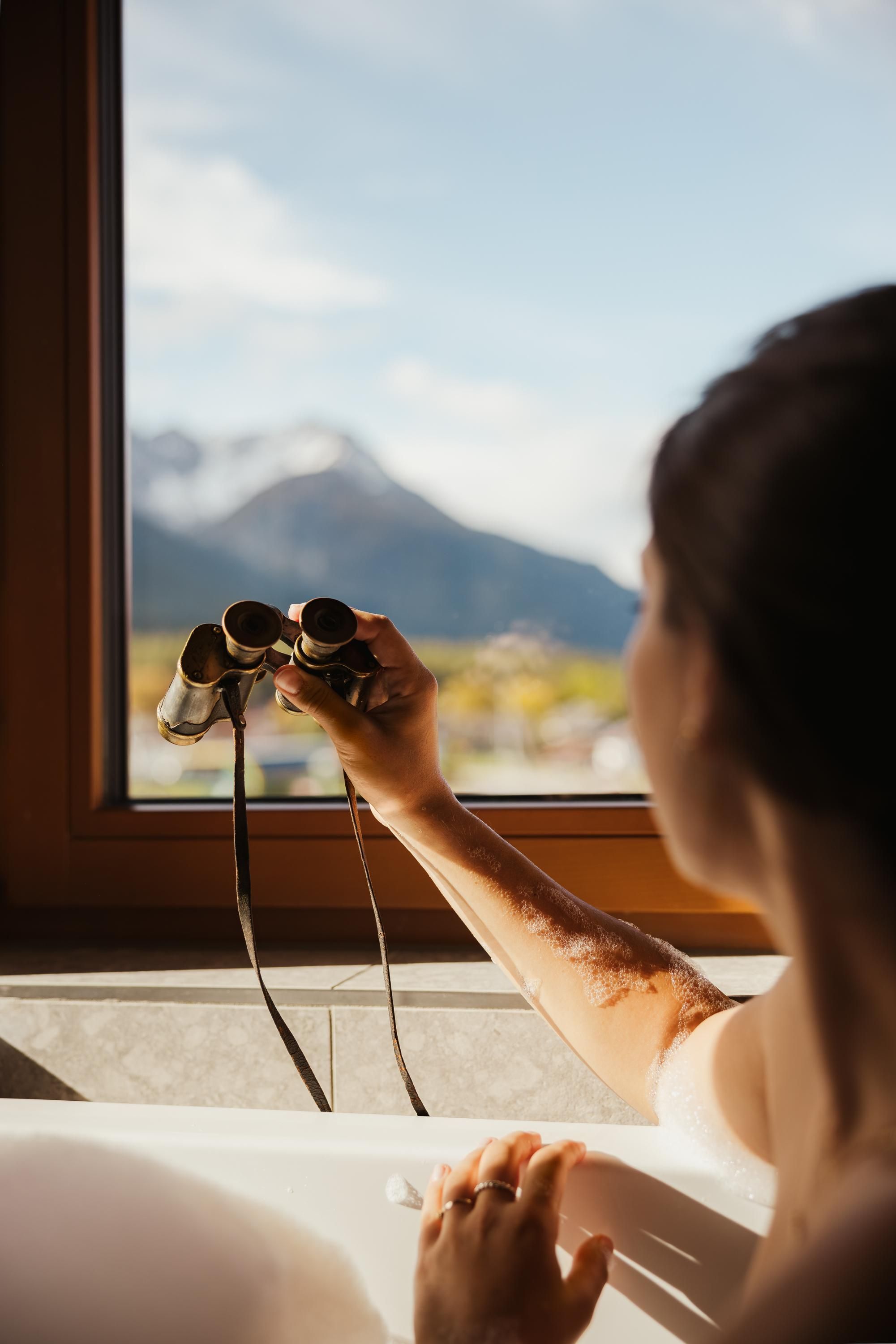 A person is looking out of a window with binoculars. In the background, there are mountains and a clear sky.
