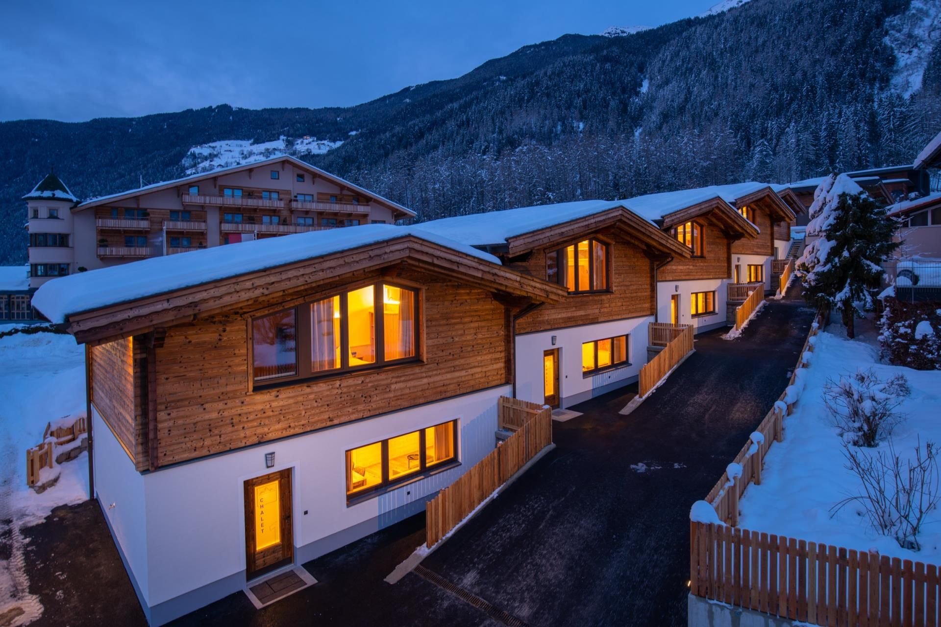 Modern wooden chalets with illuminated windows in a snowy environment. In the background, mountains and a hotel are visible.