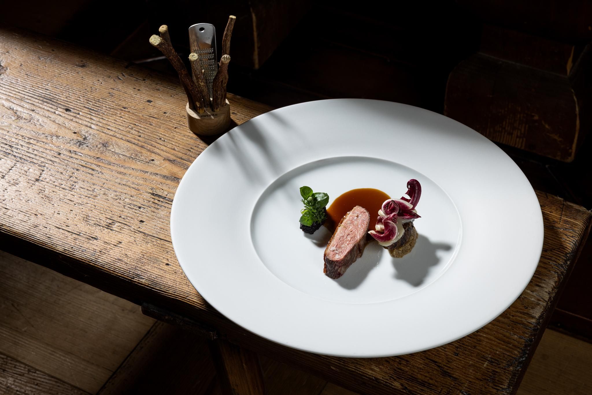 An artistically arranged plate with tender meat, colorful vegetables, and a sauce. The table is made of wood and has a rustic appearance.