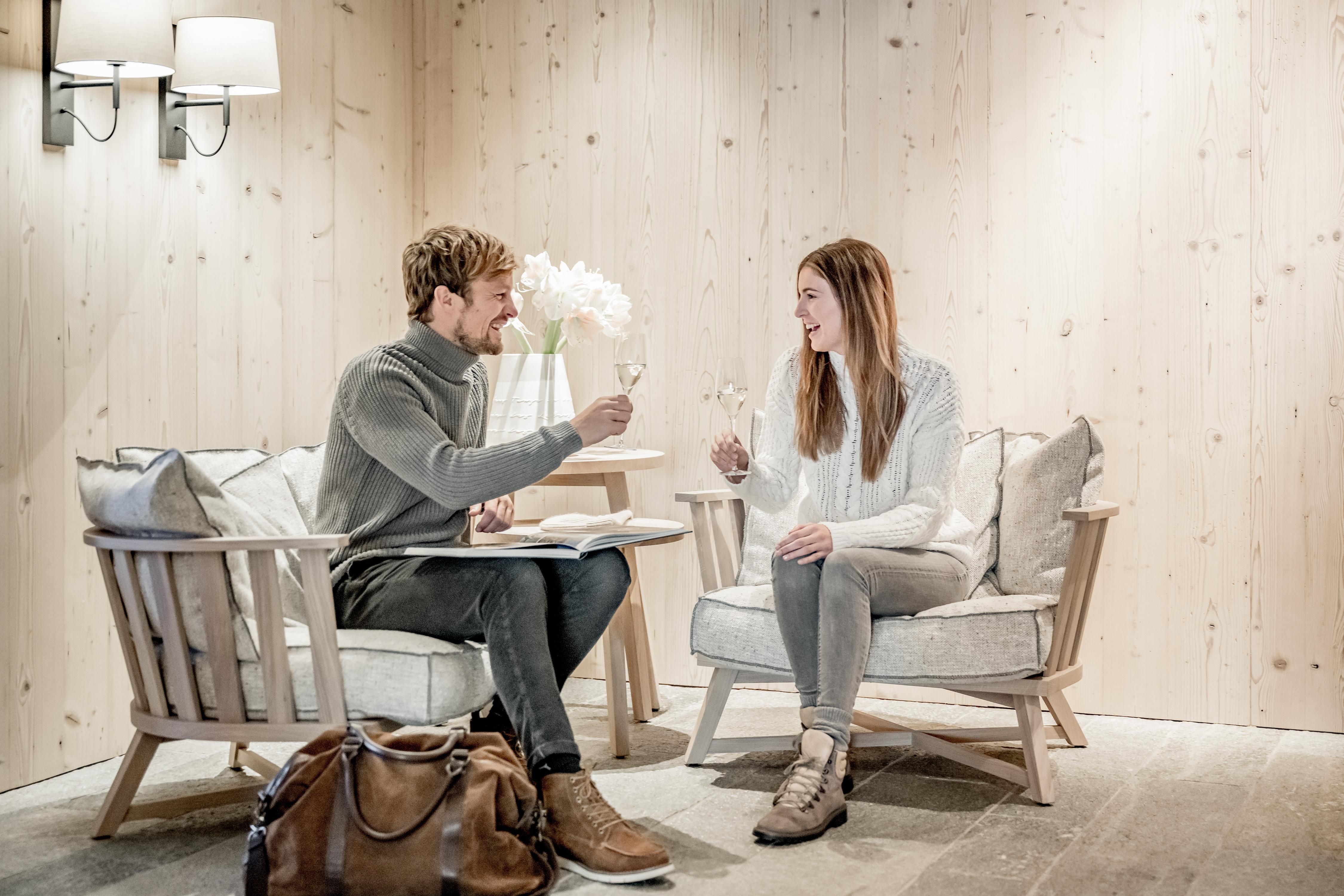 A couple sits in cozy chairs and cheers each other with glasses. The warm wooden paneling creates an inviting atmosphere.
