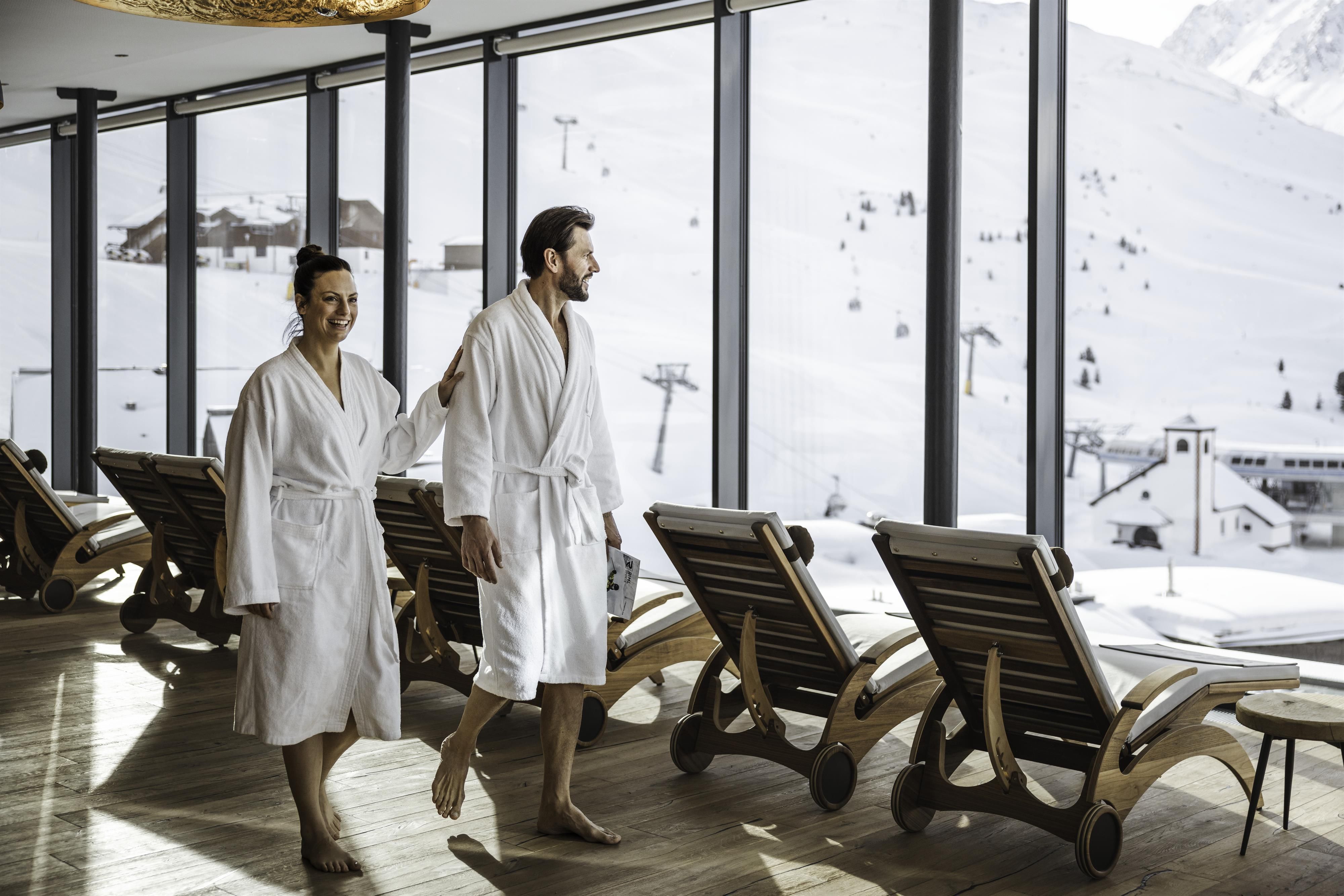 A modern spa with large windows offering views of snow-covered mountains. Two people in bathrobes are enjoying the relaxing atmosphere.
