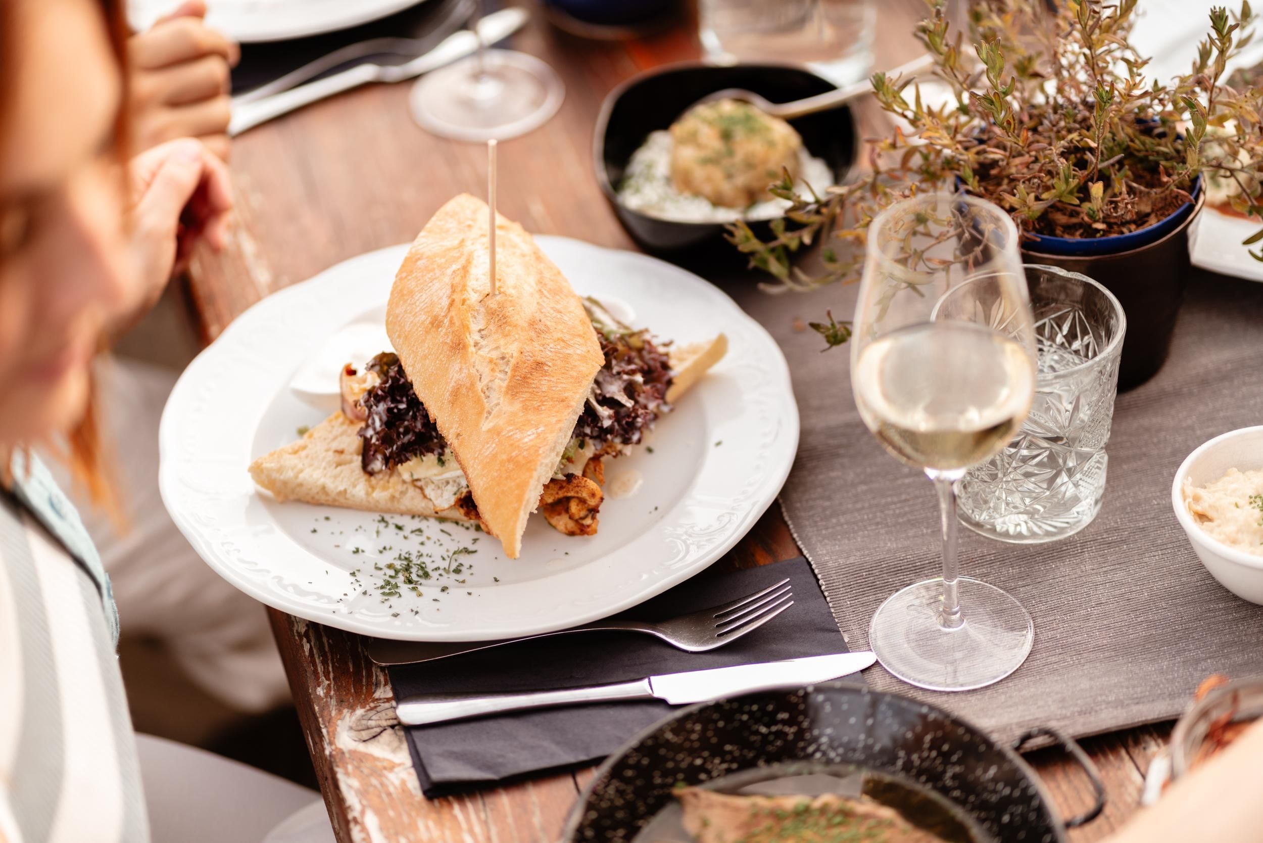 A delicious dish on a white plate, consisting of bread and meat, is arranged on a nicely set table. In the background, there is a glass of white wine and small bowls with side dishes.