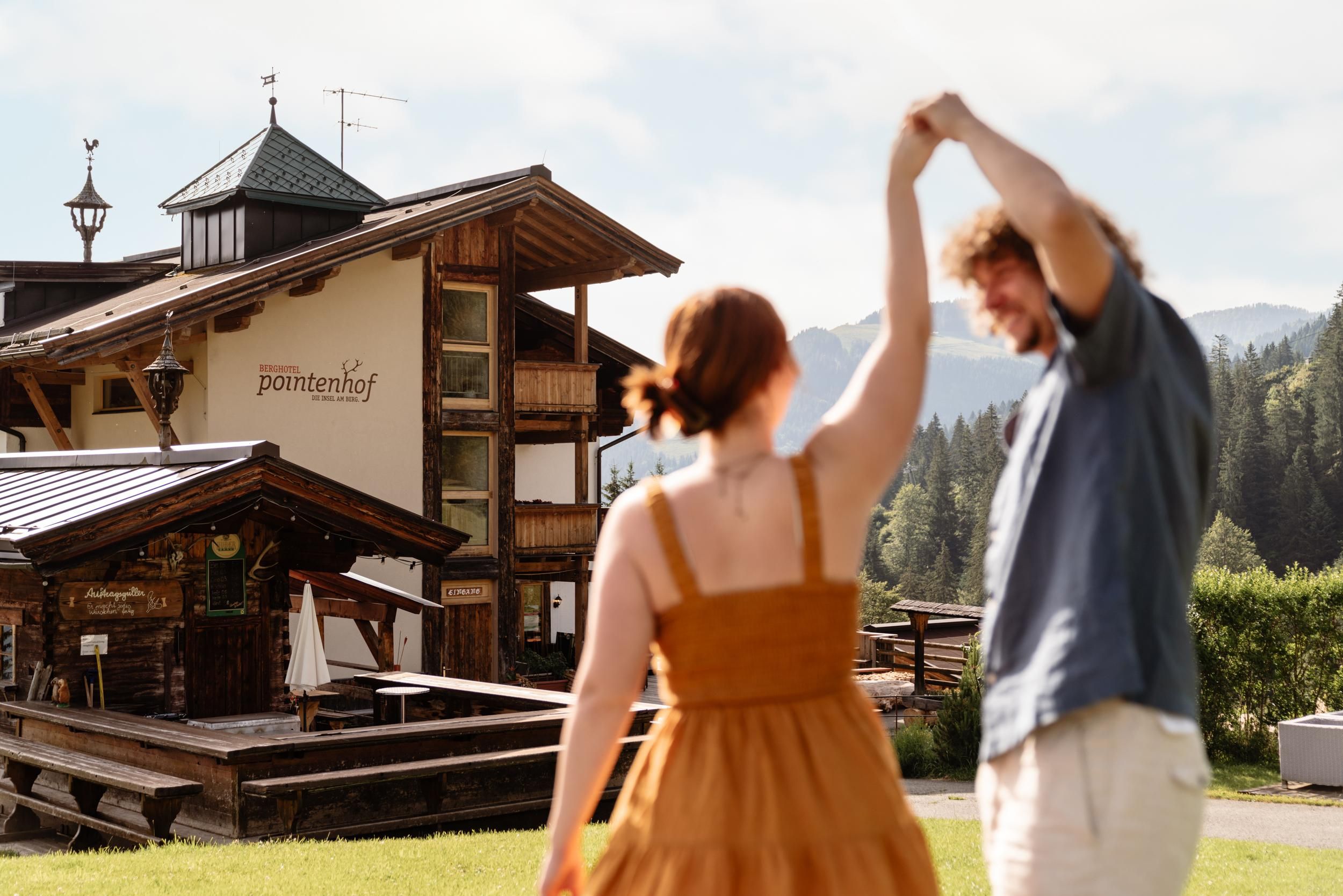 A couple dances happily outdoors in front of a wooden building. In the background, trees and mountains can be seen.