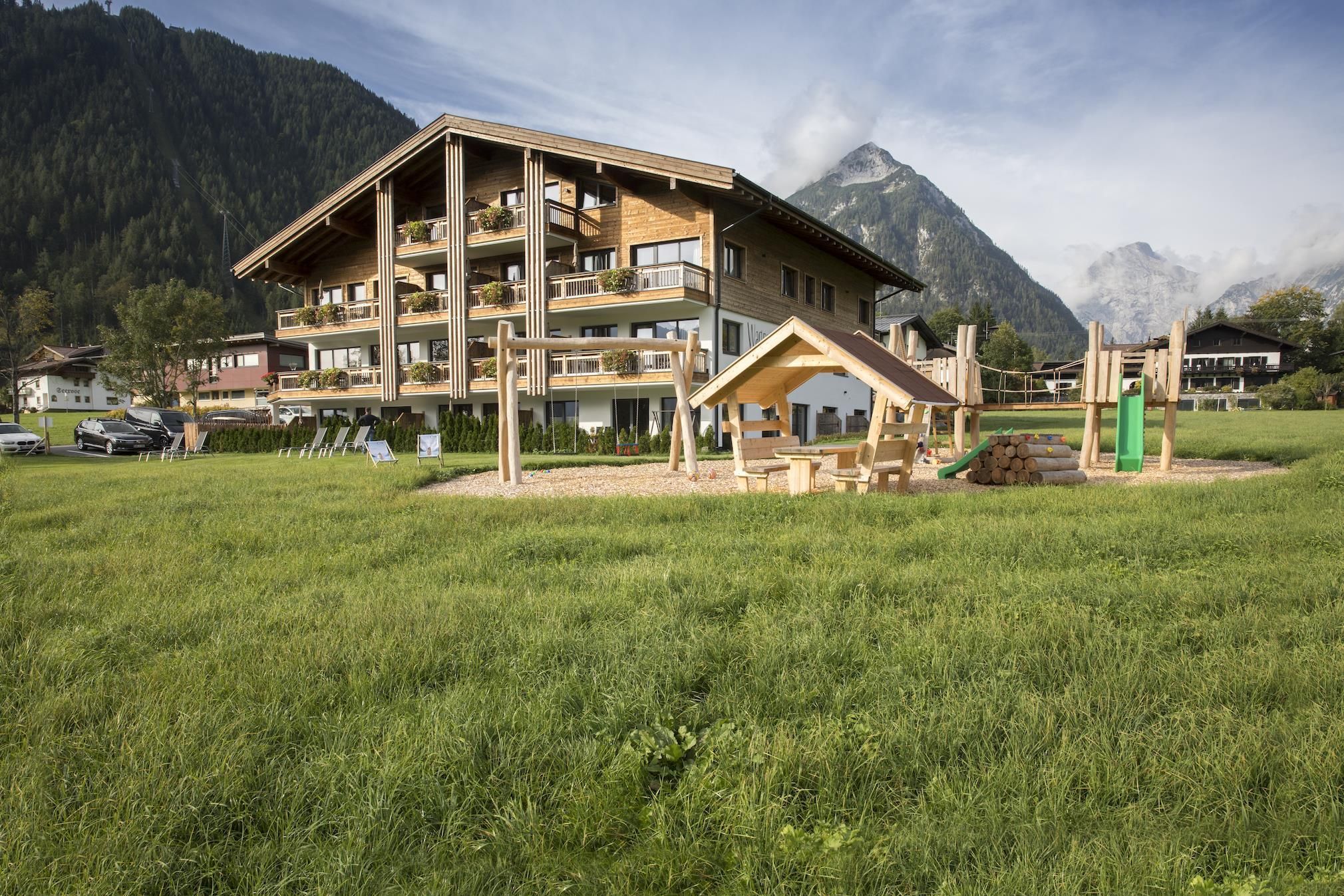 A modern building with balconies amidst a green meadow. In the foreground, there is a playground with wooden structures and a slide.
