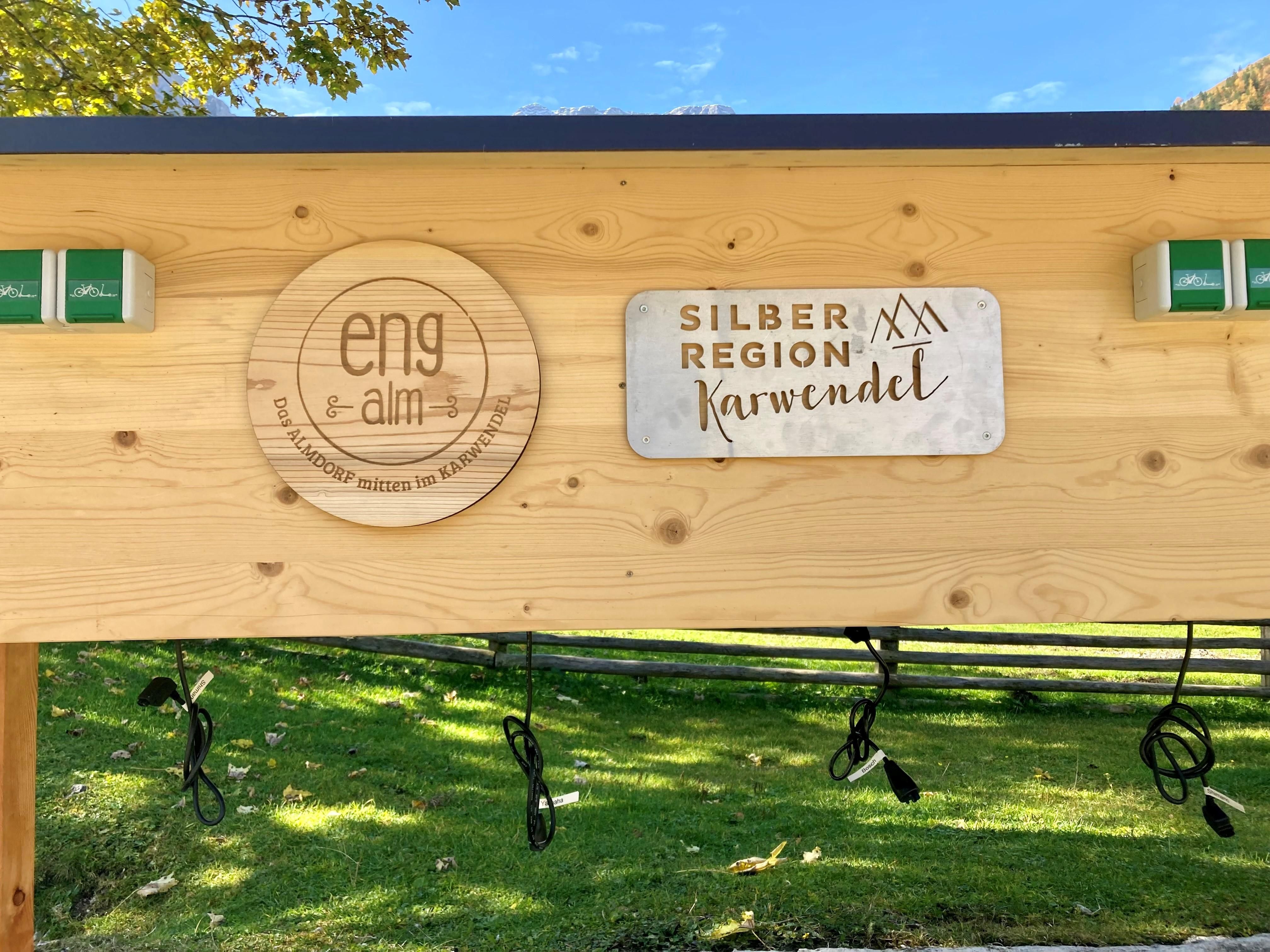 A wooden sign with the logos of "eng alm" and "Silberregion Karwendel". It stands in a green environment under a blue sky.