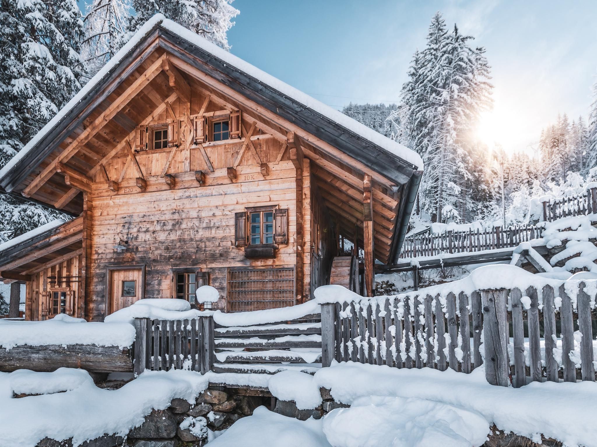 An idyllic wooden house in the snow, surrounded by a picturesque garden. The sun is shining behind the trees, creating a cozy atmosphere.