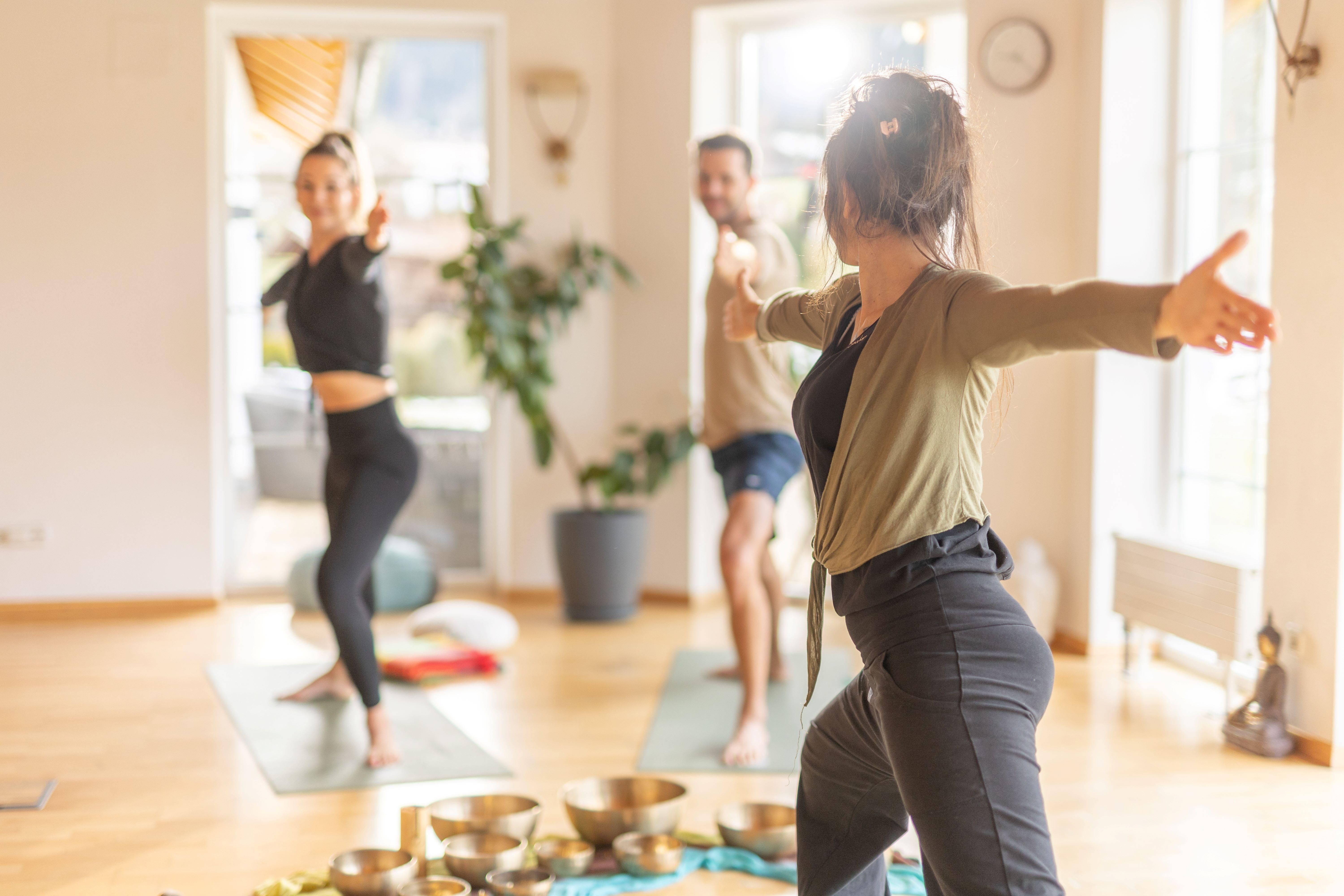 A group of people is practicing yoga in a bright room. In the foreground, there are singing bowls on a colorful cloth.