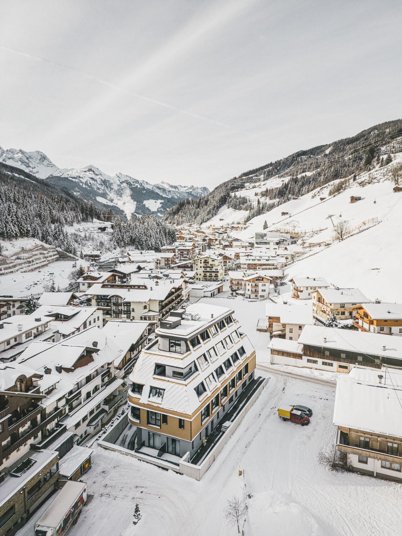 A snowy mountain landscape with a charming village. In the foreground stands a modern building surrounded by snow-covered roofs.