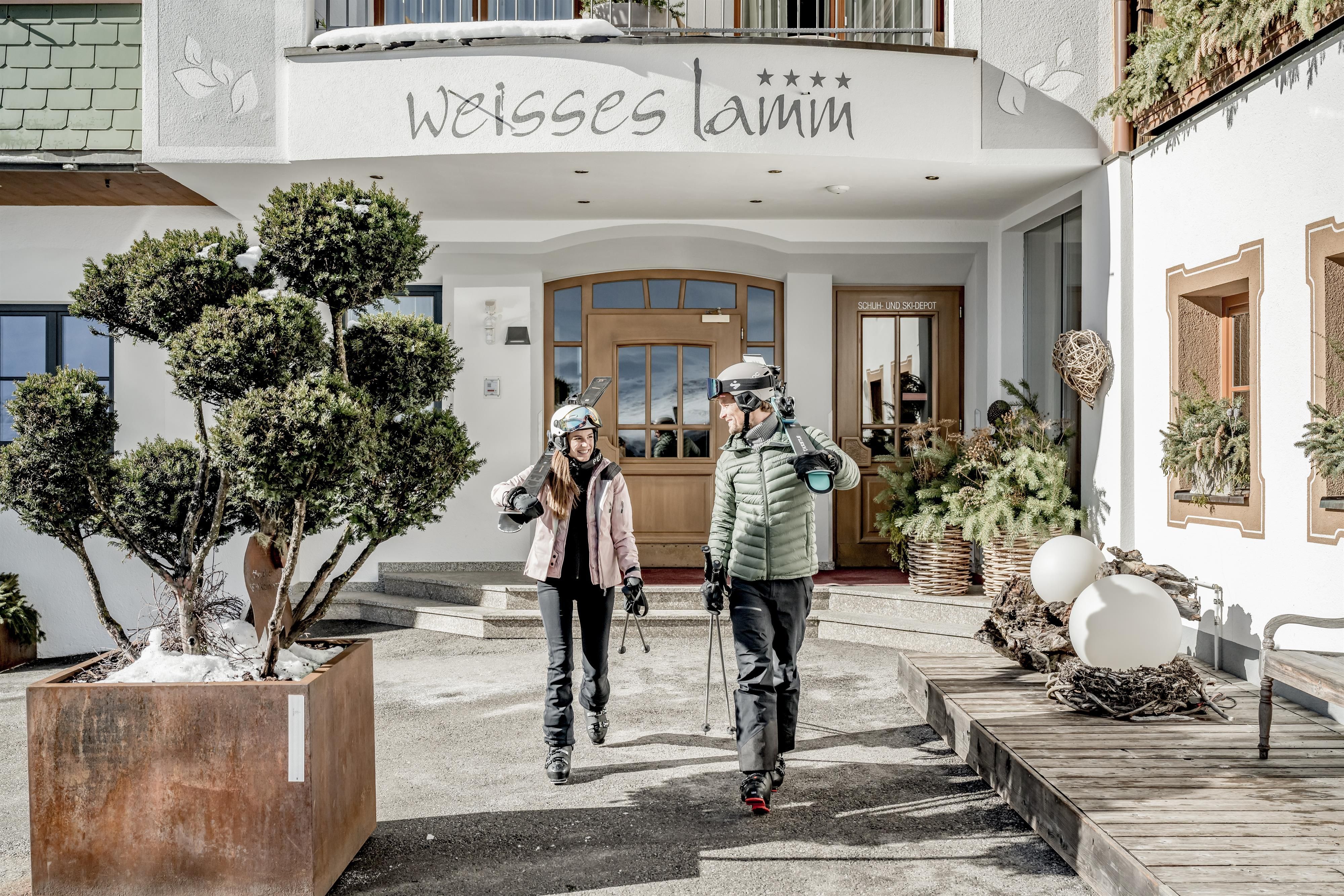 Two people are walking in winter clothing in front of a hotel named "white lamb." In the background, plants and the entrance area of the hotel are visible.
