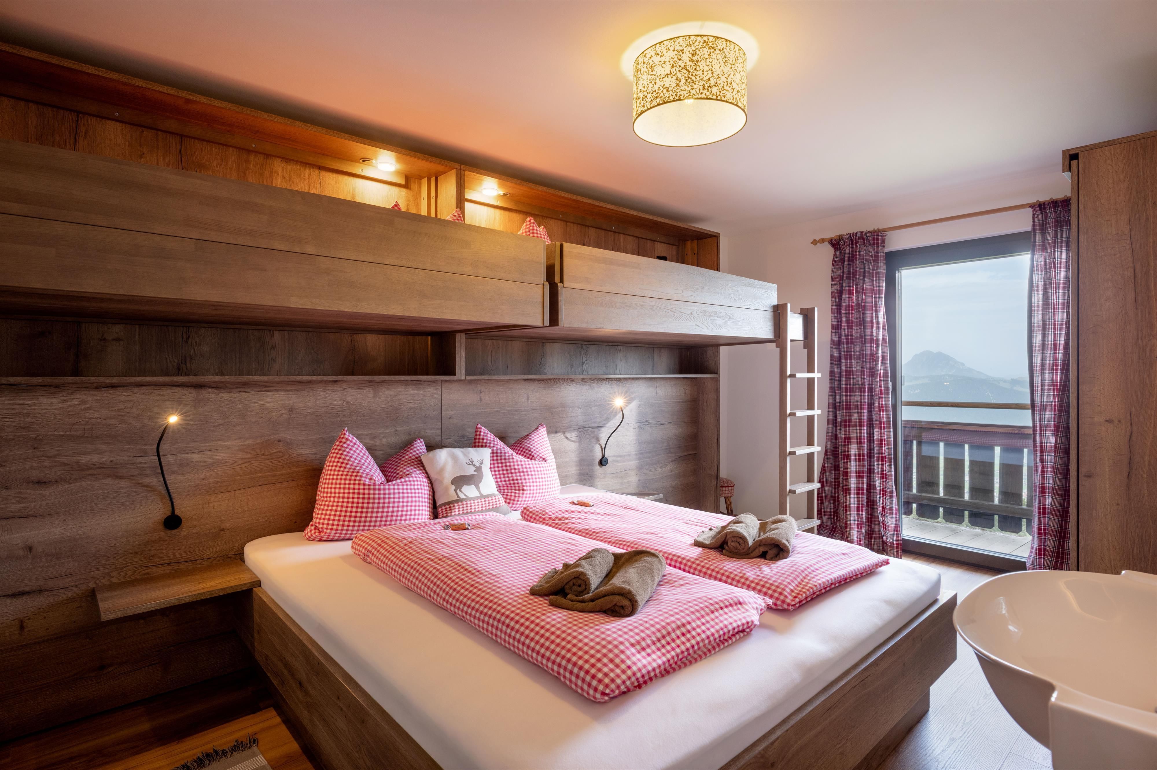A modern bedroom with wooden interior and a large window. The bedding is in soft pink tones.