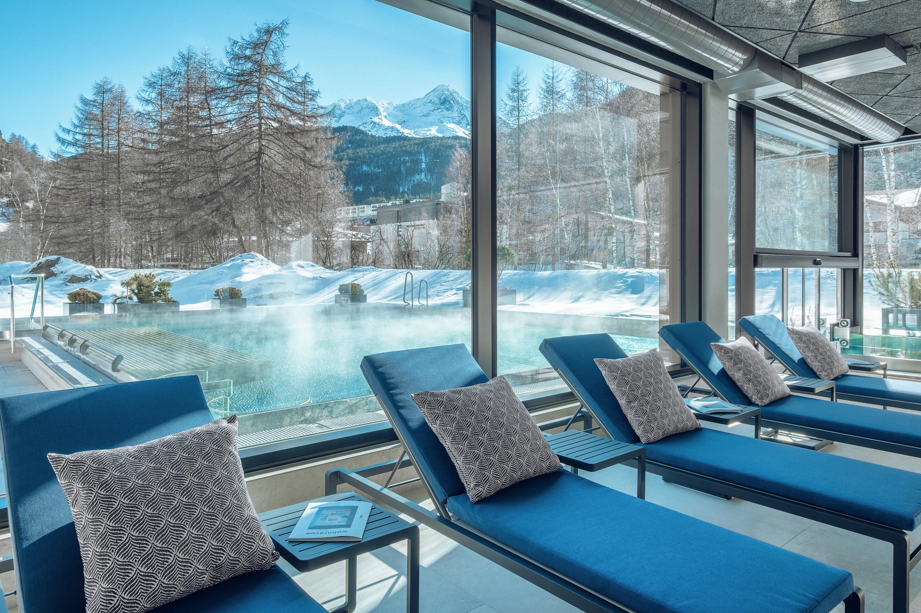 A modern wellness area with loungers and a view of a snowy landscape. Outside, mountains and a tranquil outdoor pool can be seen.