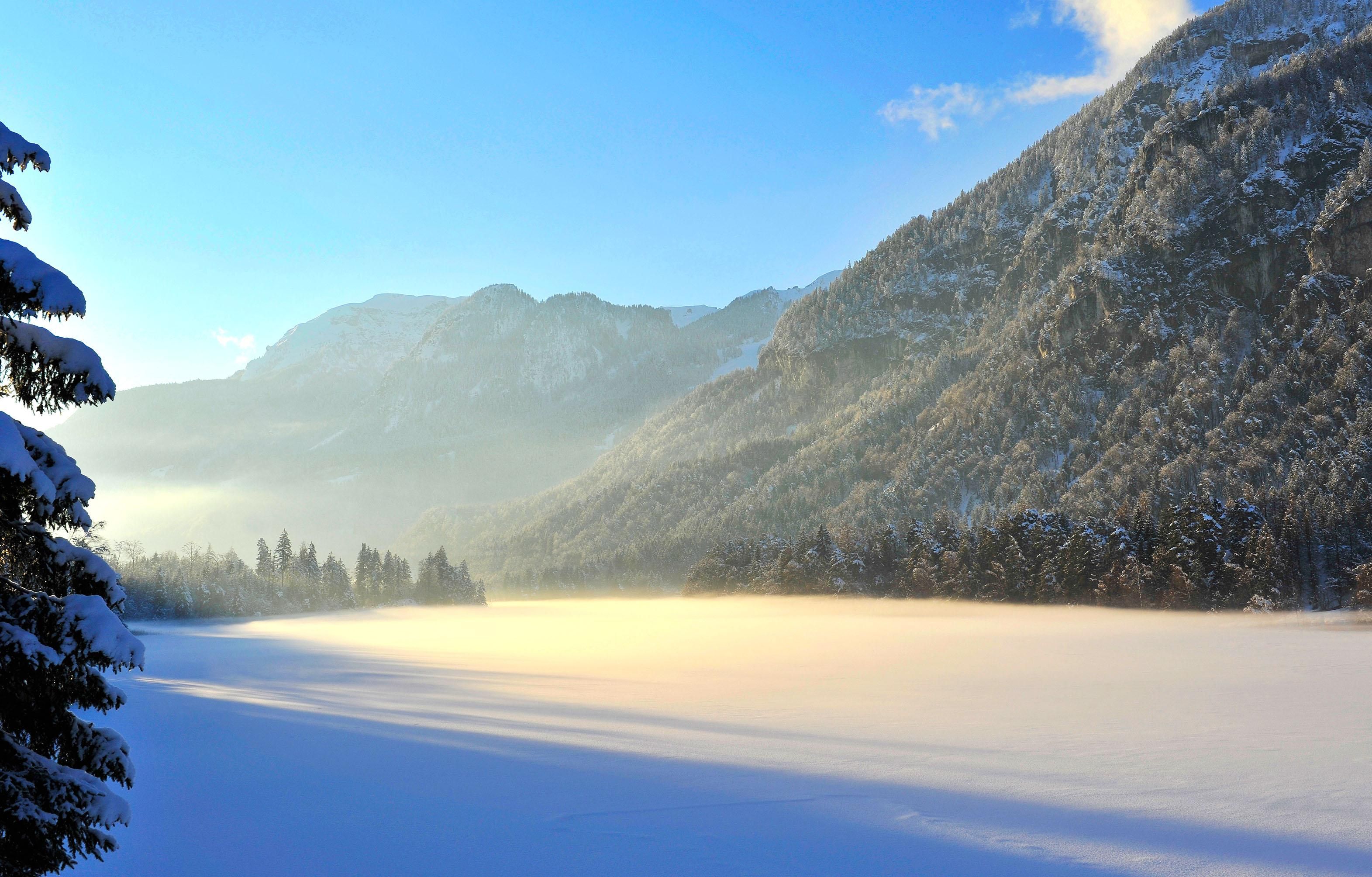 A winter landscape with snow-covered mountains and a frozen lake. The sky is clear and blue, enhancing the tranquility of the scene.