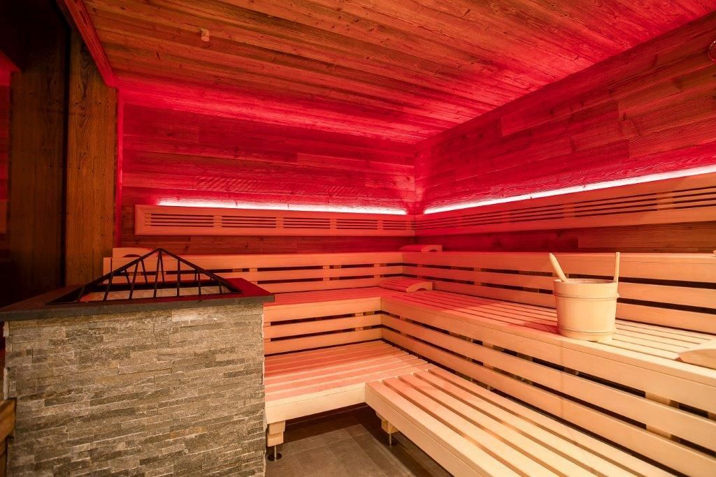 A modern sauna with wooden shelves and gentle red lighting. In the room, there is a stone fireplace and a wood container.