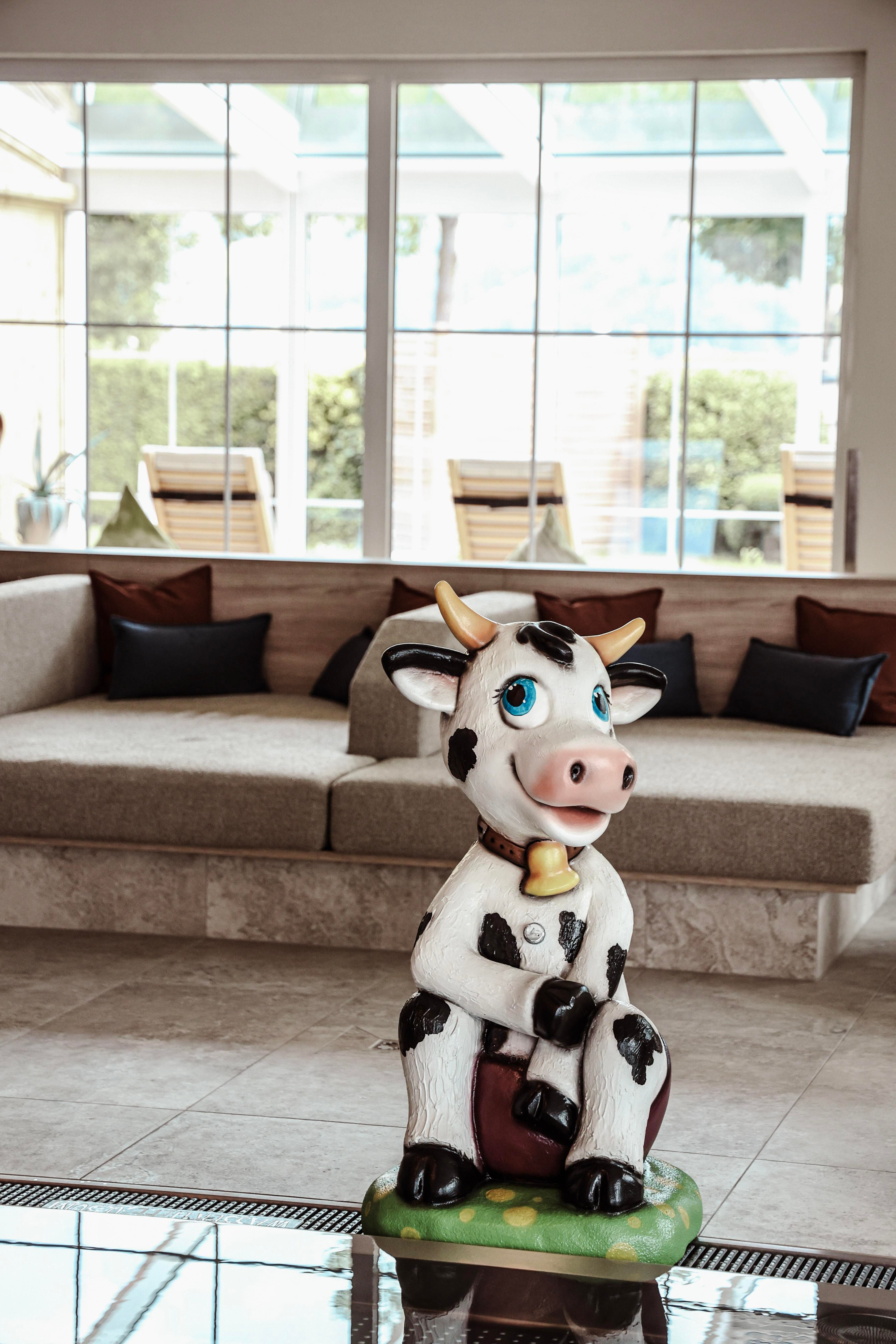 A cute cow figure sits on a stool in a modern room. In the background, windows and a cozy couch are visible.