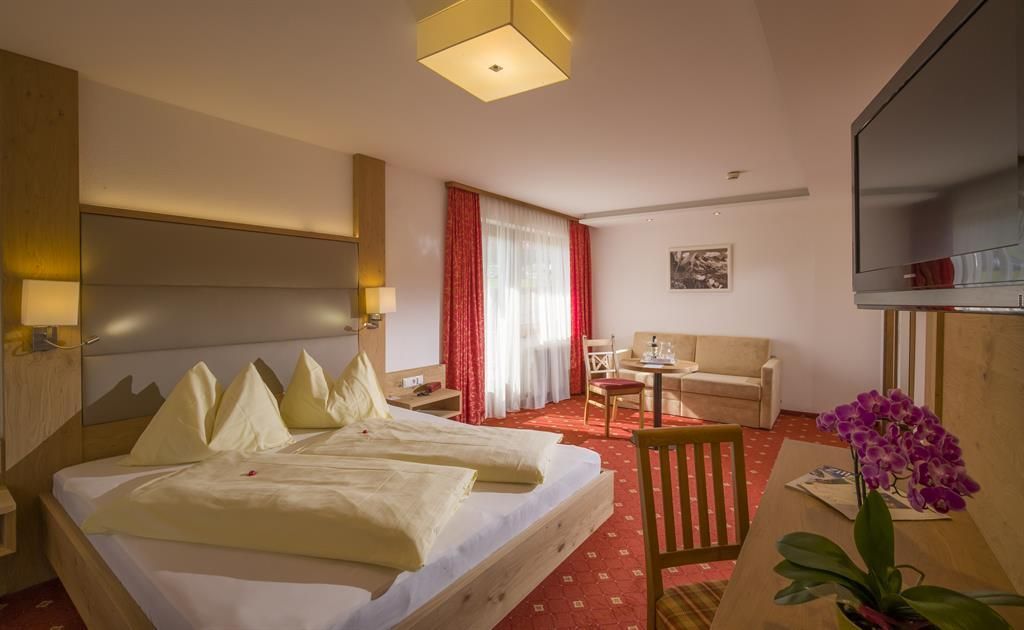 A modern hotel room with a large double bed and cozy furniture. Plenty of light streams through the window, and a plant sits on the table.