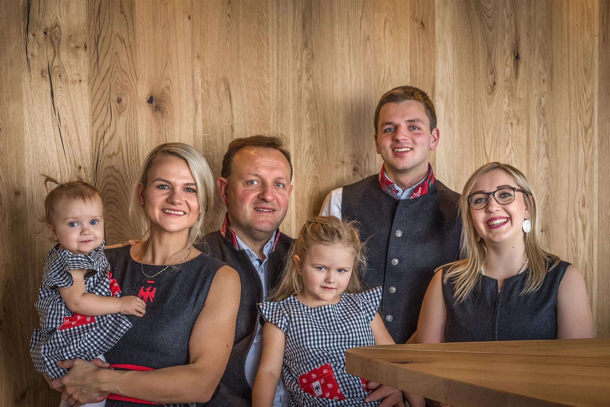 A family is posing in front of a wooden-paneled wall.
Everyone is wearing traditionally inspired clothing and has friendly faces.