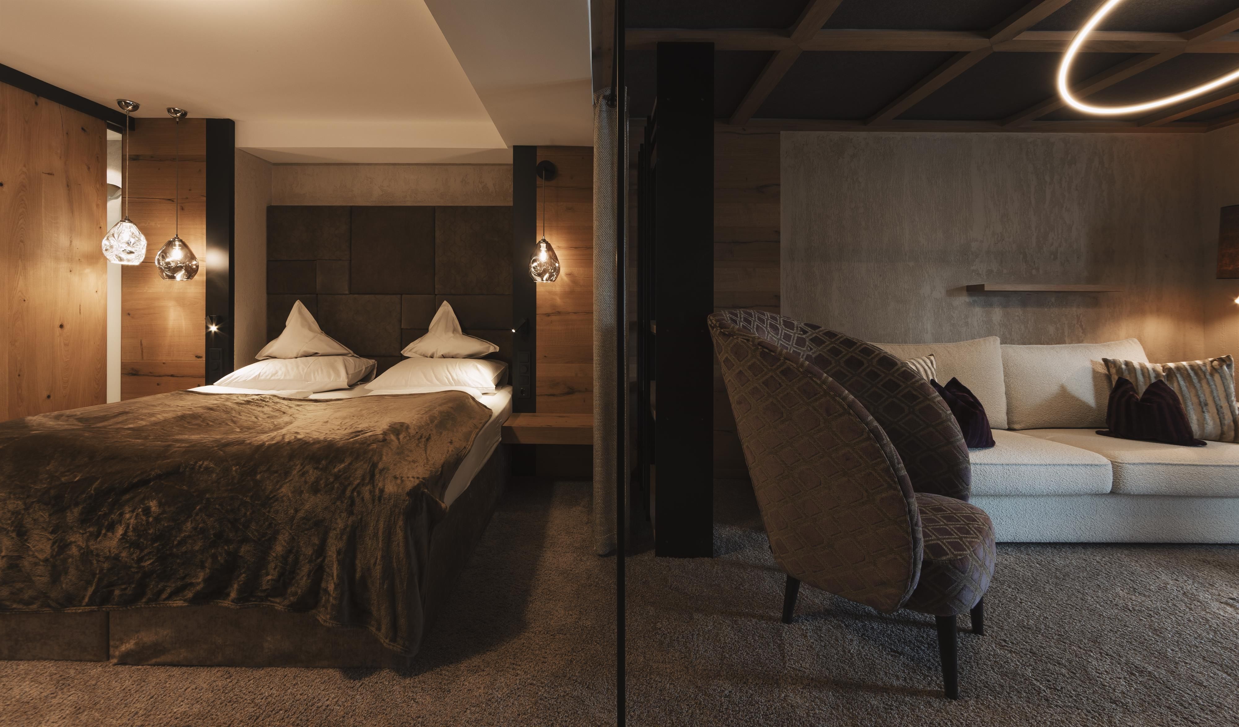 A modern bedroom with a large bed and cozy wooden elements. On the other side, there is a stylish seating area with an elegant armchair and a sofa landscape.