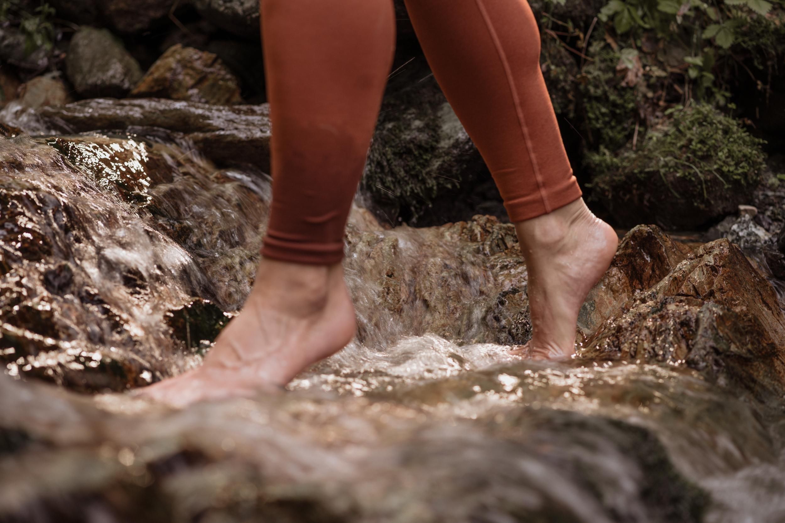 A person with bare feet walks through a small, bubbling body of water. Surrounded by stones and plants, the scene conveys a natural and soothing atmosphere.