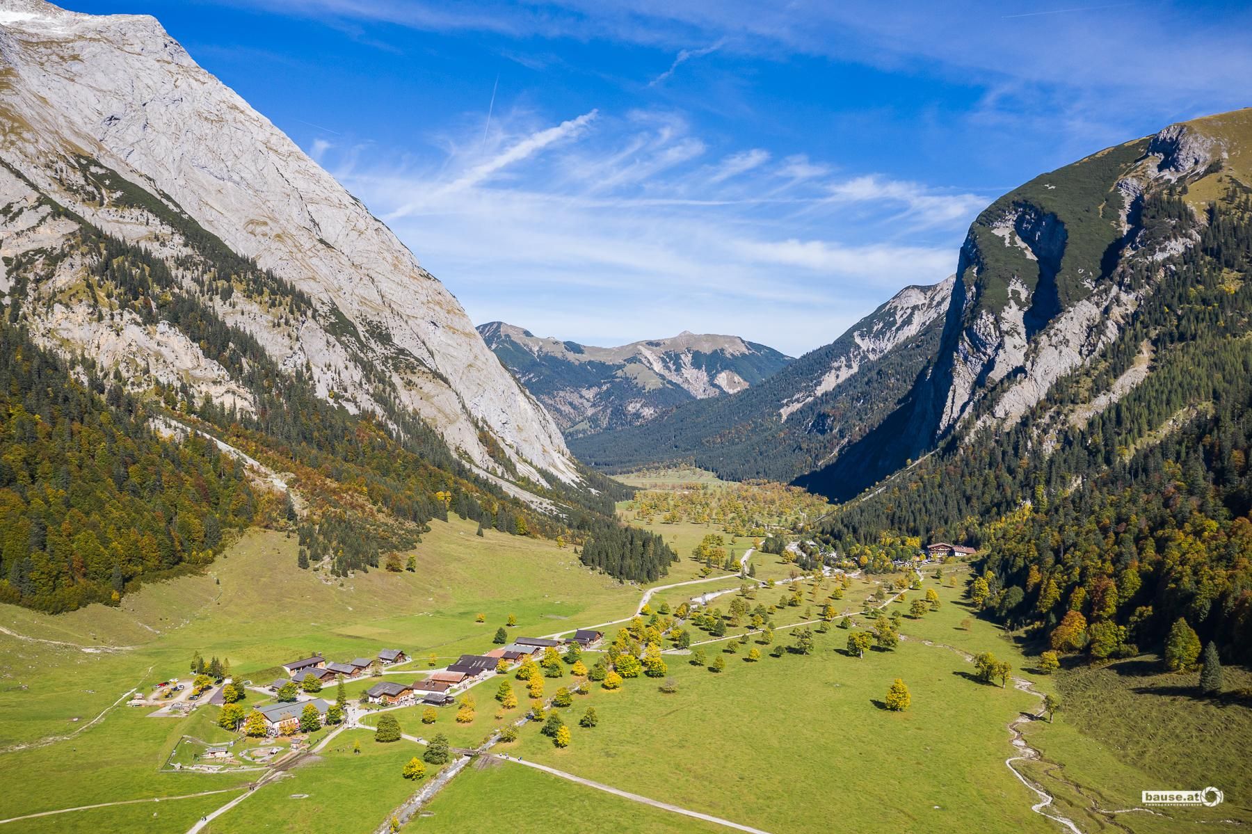 A picturesque mountain landscape with high peaks and a green valley. In the valley, small buildings and trees are scattered.