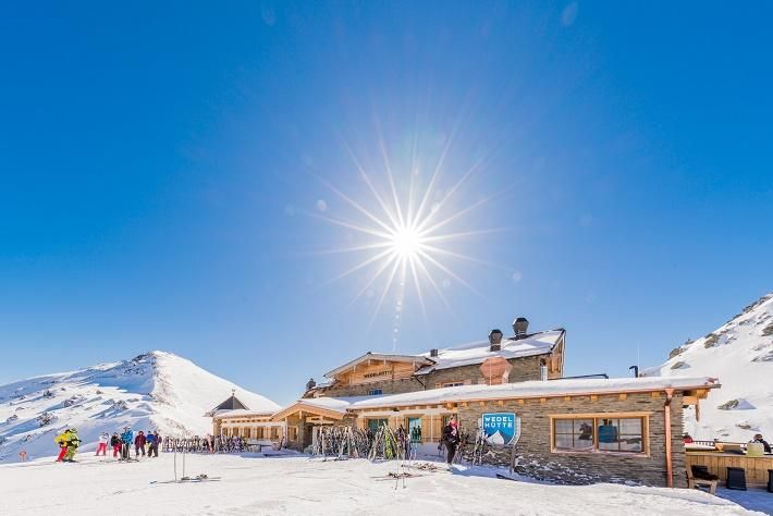 A mountain house in a snow-covered landscape under a clear blue sky. Sun rays shine brightly on the building and the skiers in front of it.
