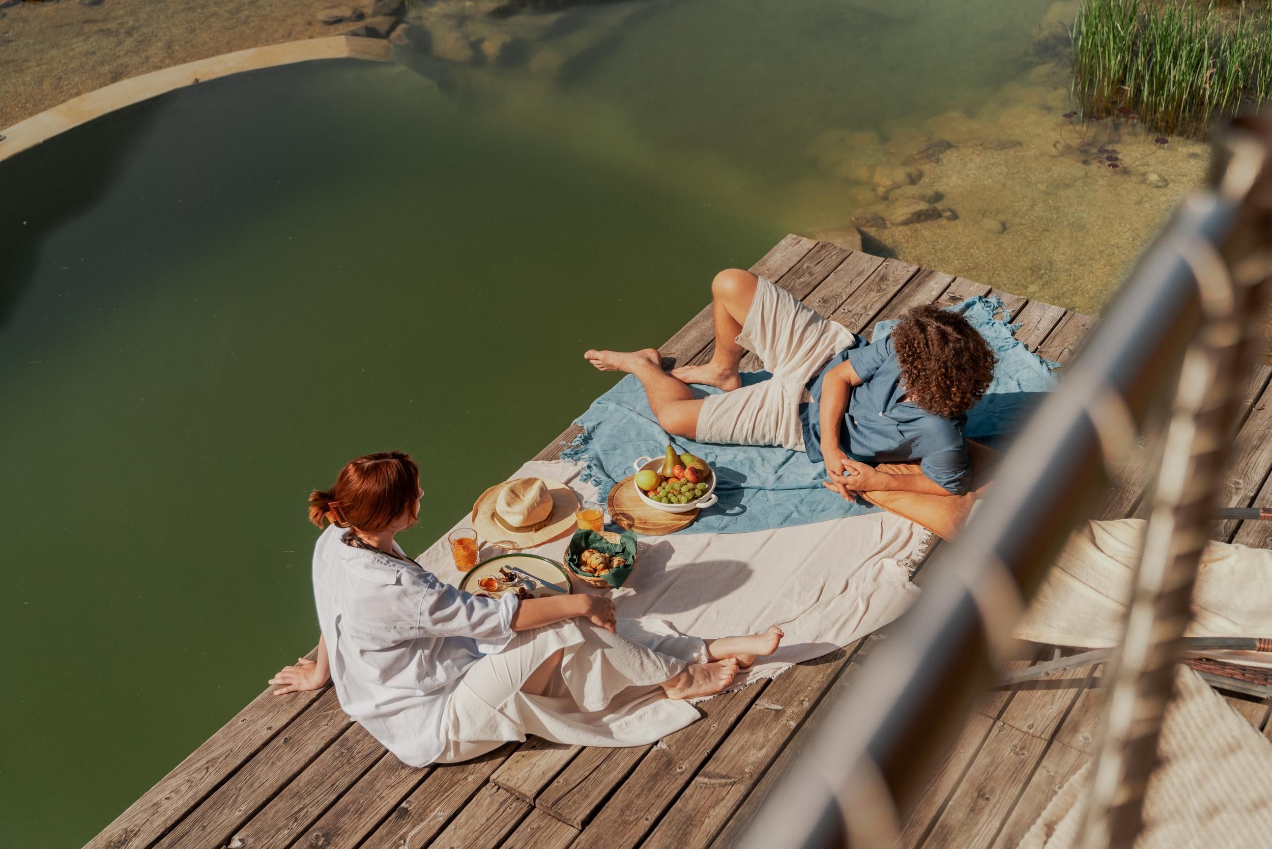 A couple enjoys a picnic by the shore of a calm body of water. They are sitting on a blanket and have a selection of fresh foods in front of them.
