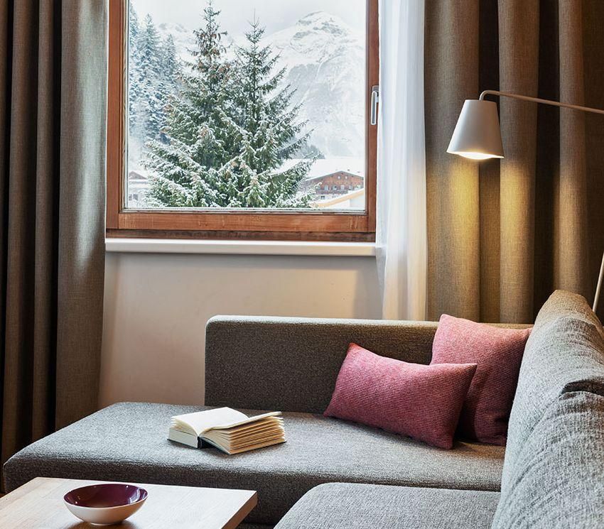 A cozy living space with a gray couch and a small table. Through the window, you can see snow-covered fir trees and mountains.