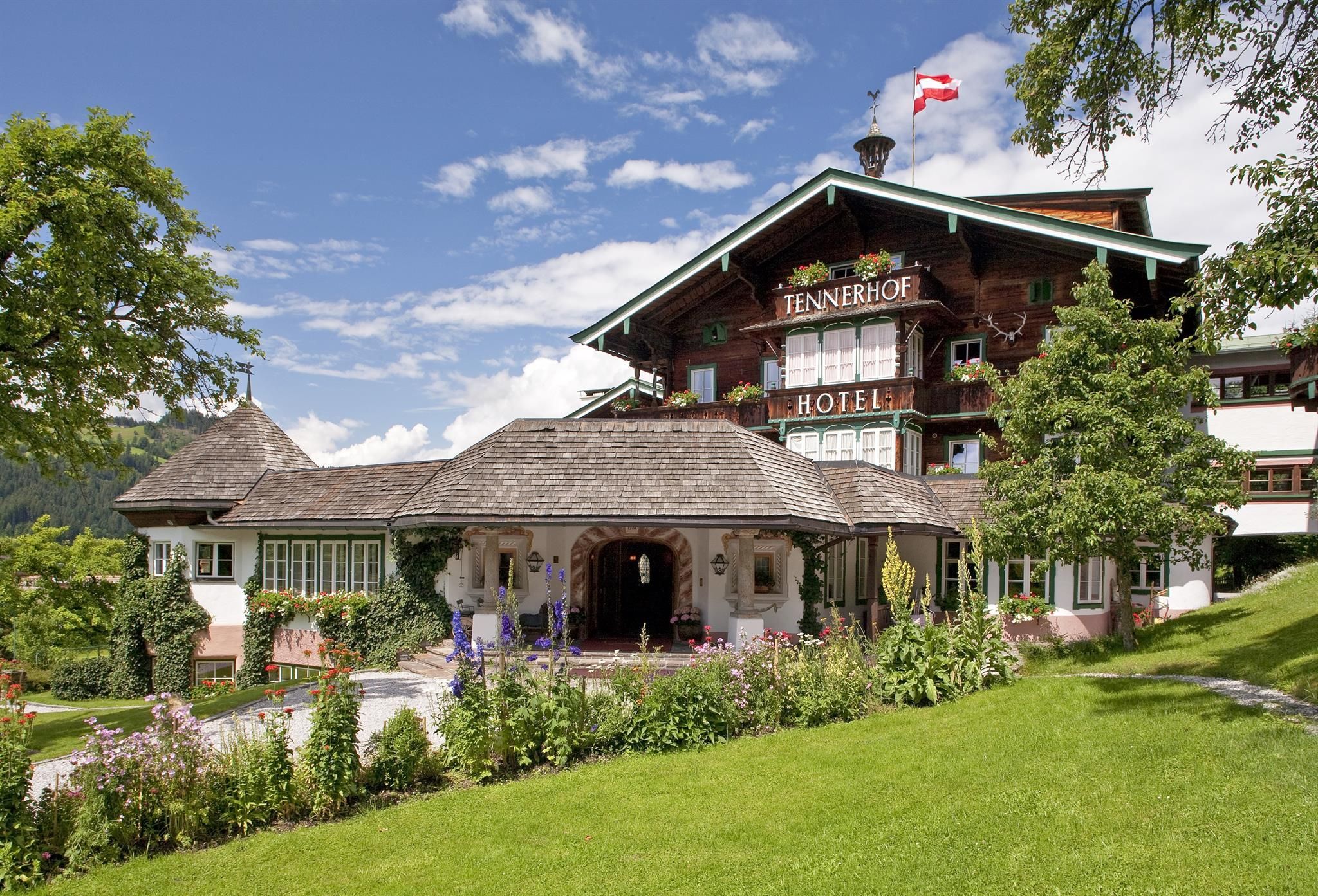 A charming hotel with a wooden facade and green meadows. Surrounded by trees and a clear blue sky.