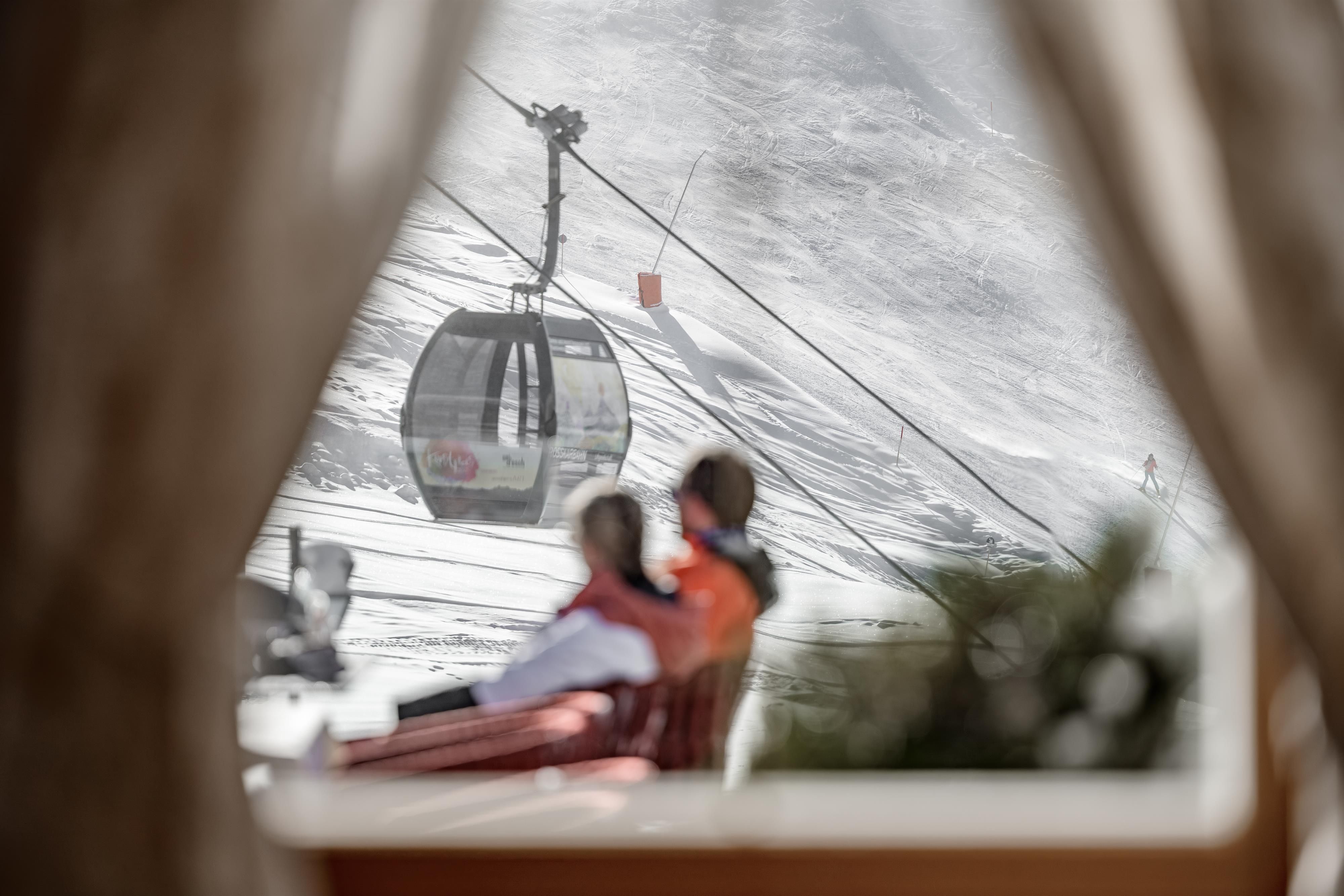 A cozy scene with two people sitting on a terrace and enjoying the view. In the background, a cable car and snowy landscape can be seen.