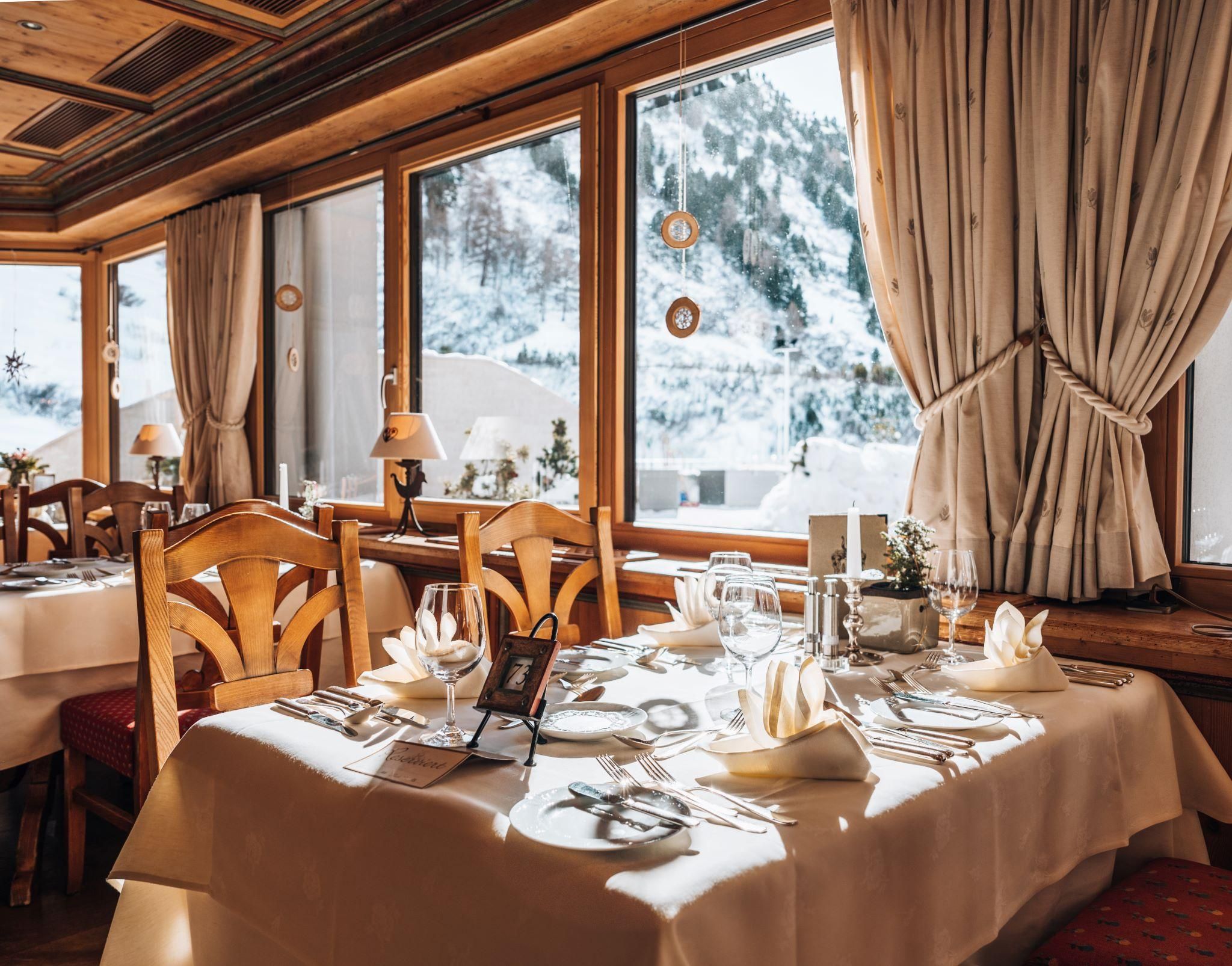A cozy restaurant with wooden furniture and elegant table decorations. The snowy mountain landscape is visible through large windows.