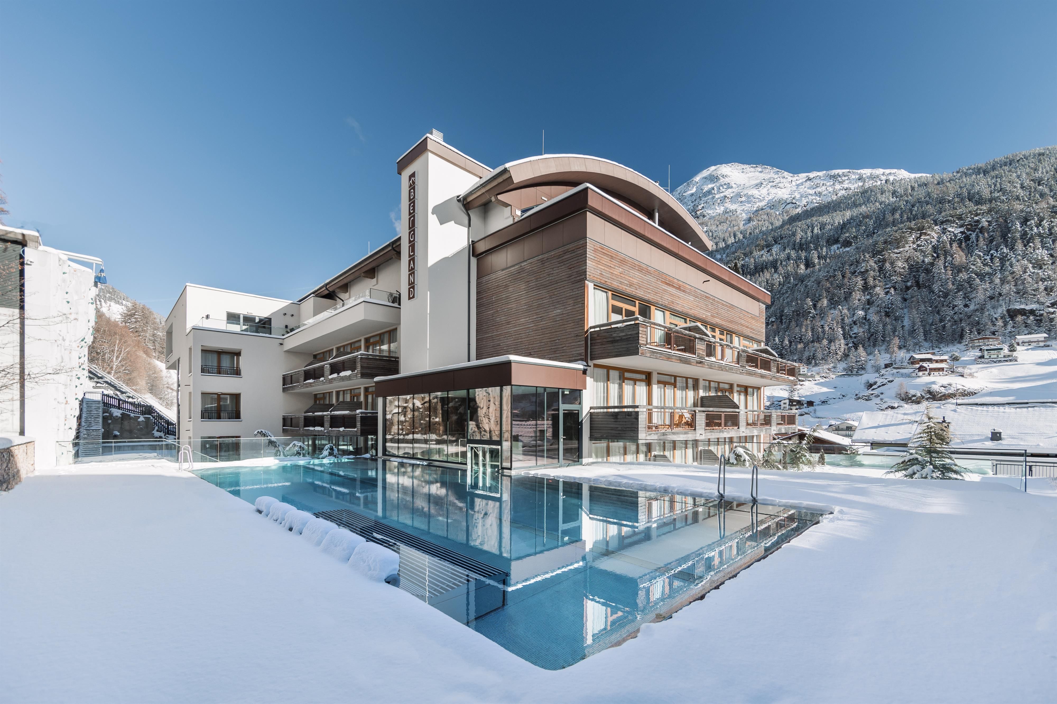 A modern hotel building in the snow with a pool in the foreground. In the background, snow-covered mountains are visible.