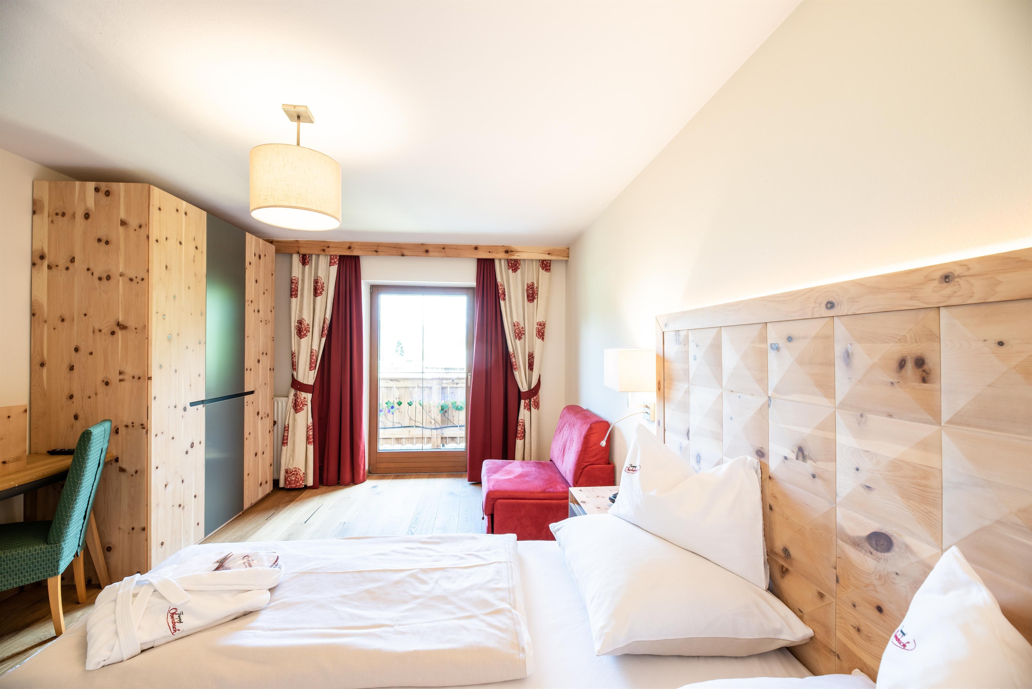 A cozy hotel room with wood-paneled walls. There is a large bed, a red armchair, and a balcony with an outside view.