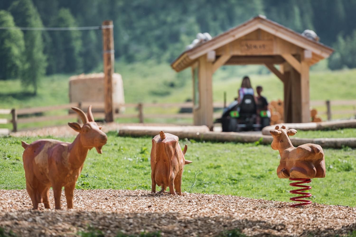 A playground scene with wooden figures of goats on a playground. In the background, there is a wooden shelter and green meadows are visible.