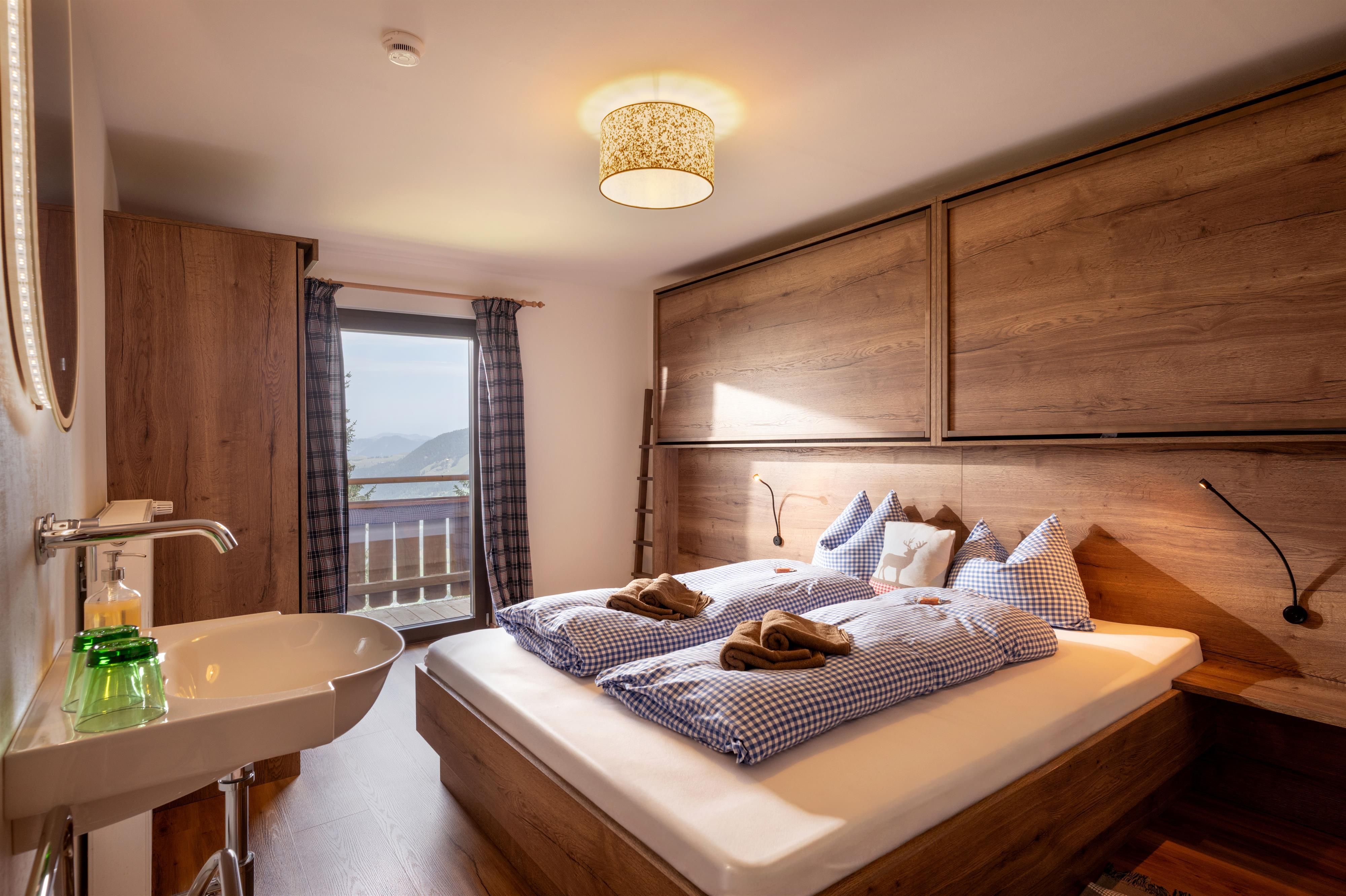 A cozy bedroom with a double bed and wooden paneling. Large windows offer a view outside and plenty of daylight.