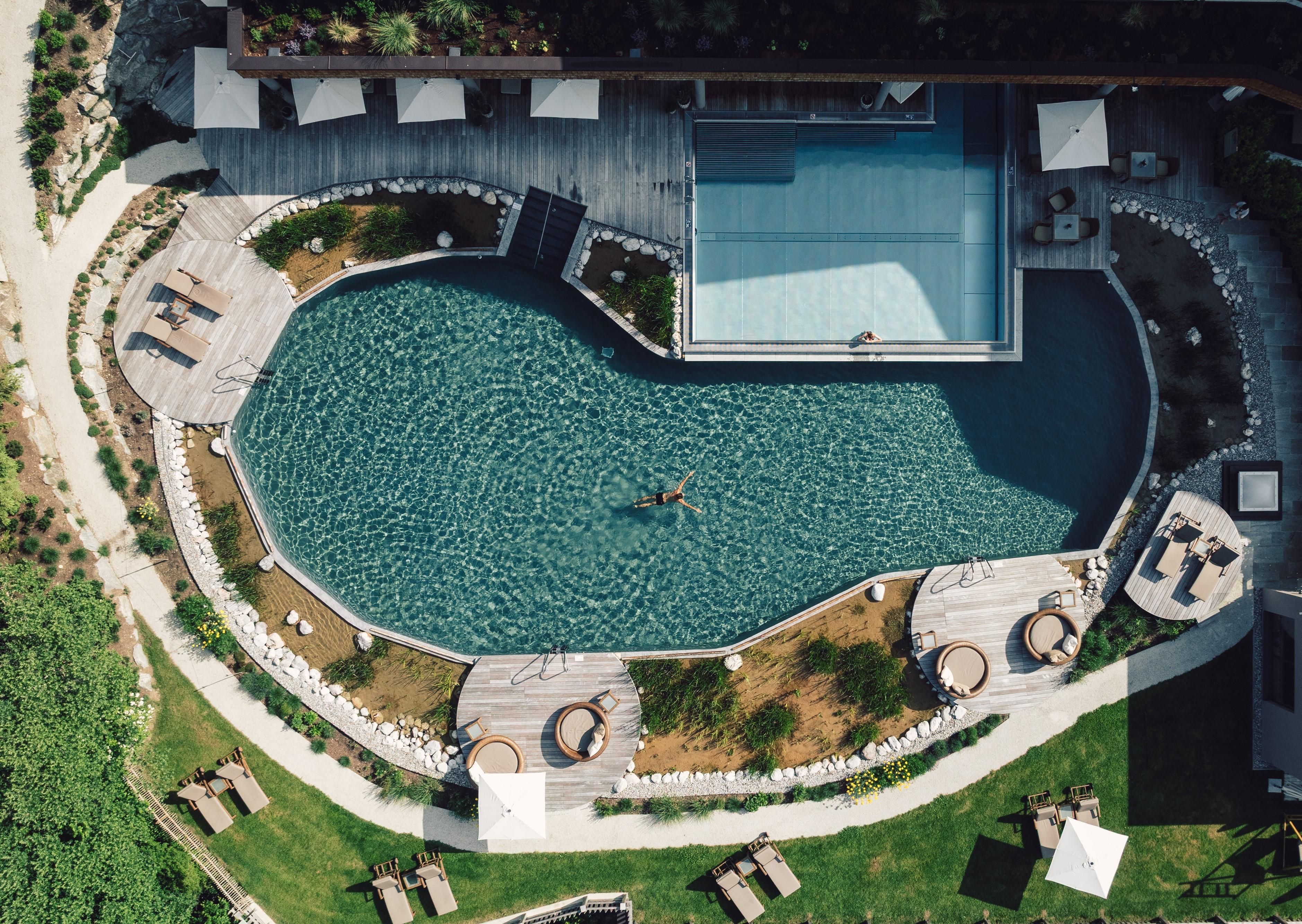 An oval pool with clear water, surrounded by wooden decks and lounge chairs. The green landscape and sun umbrellas complement the relaxing atmosphere.