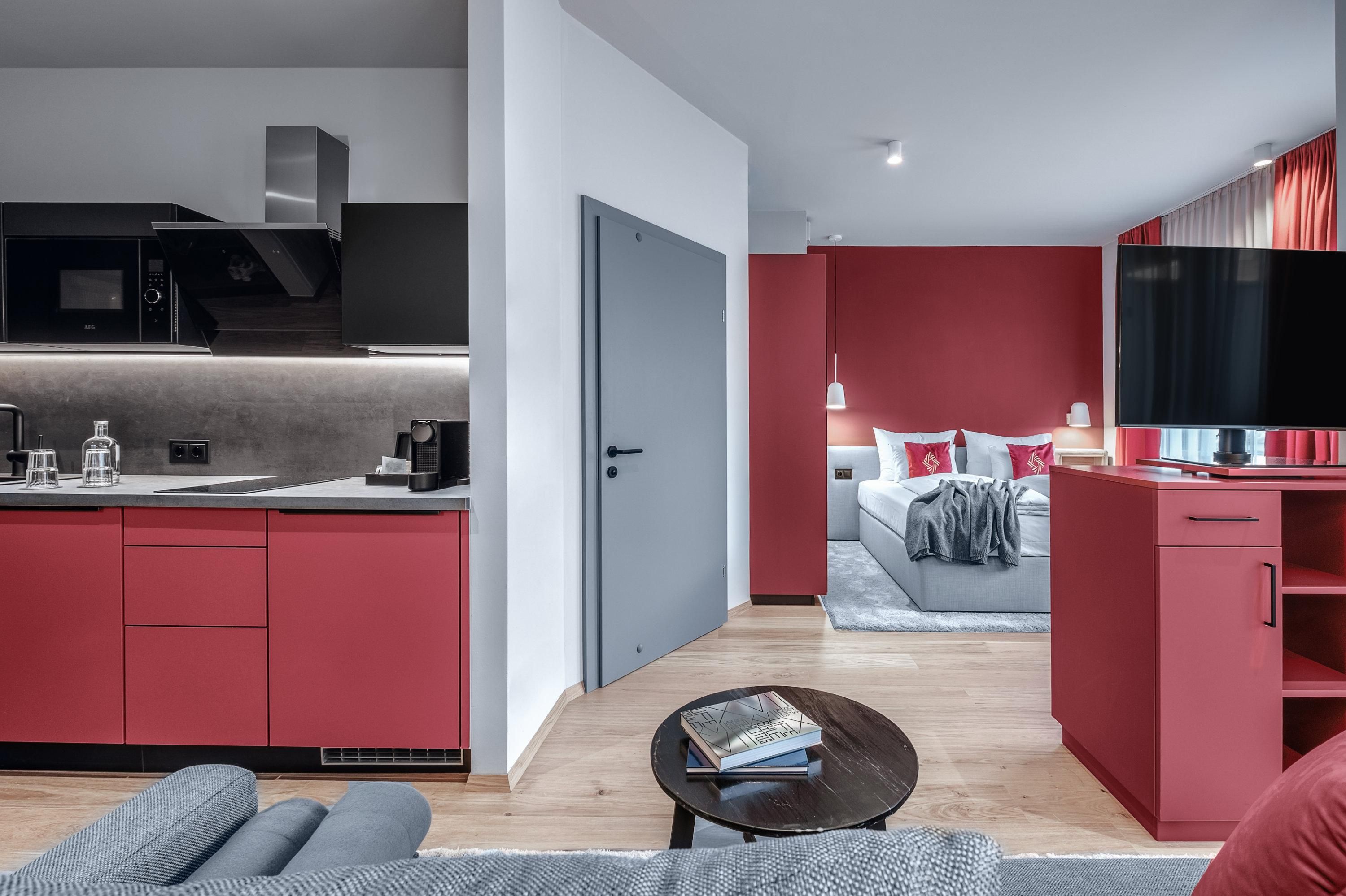 A modern living kitchen with red accents and elegant furnishings. In the background, a cozy bedroom with a large bed is visible.