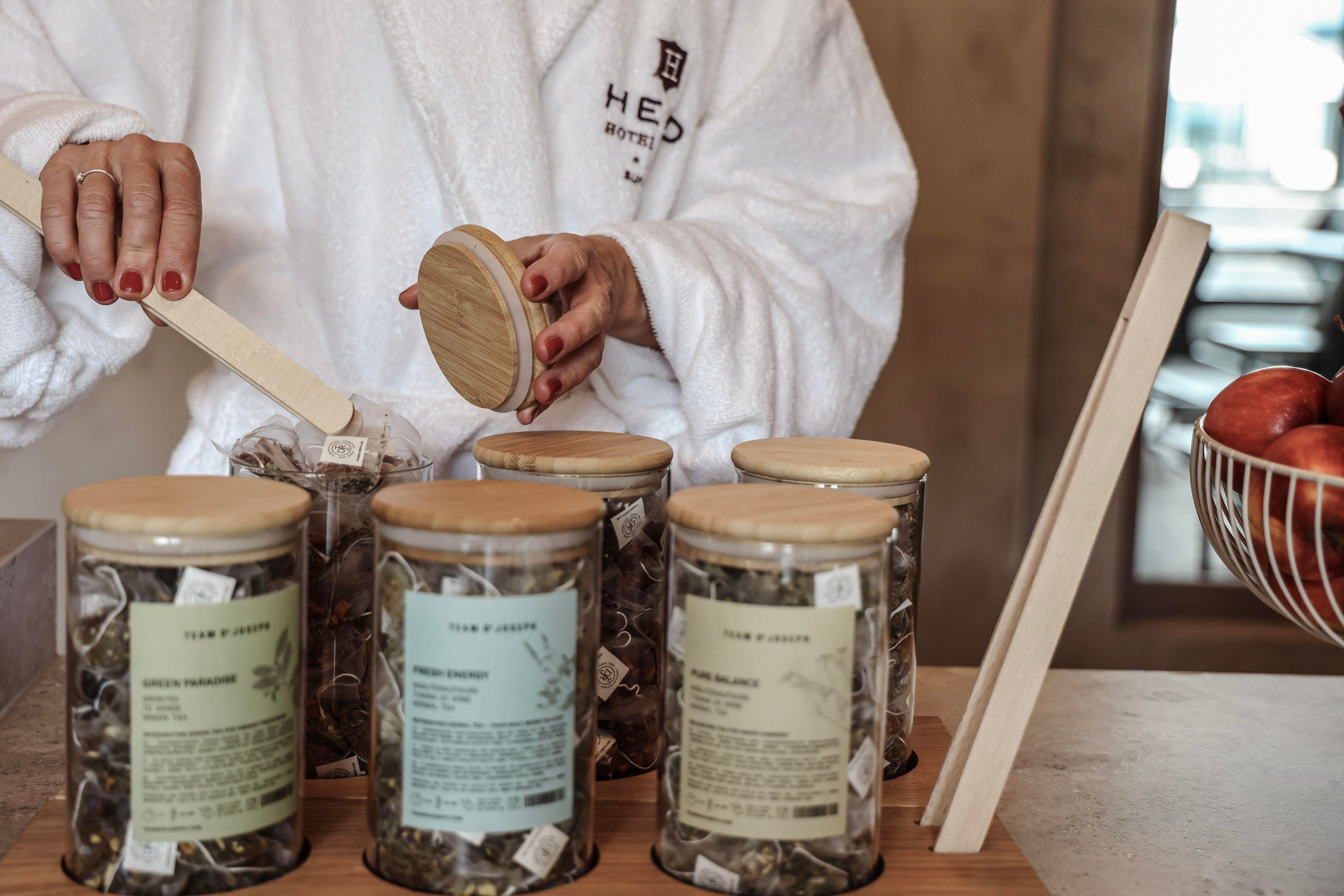 A person in a bathrobe is taking a tea bag out of a glass container. In the background, more tea glasses and a fruit bowl can be seen.