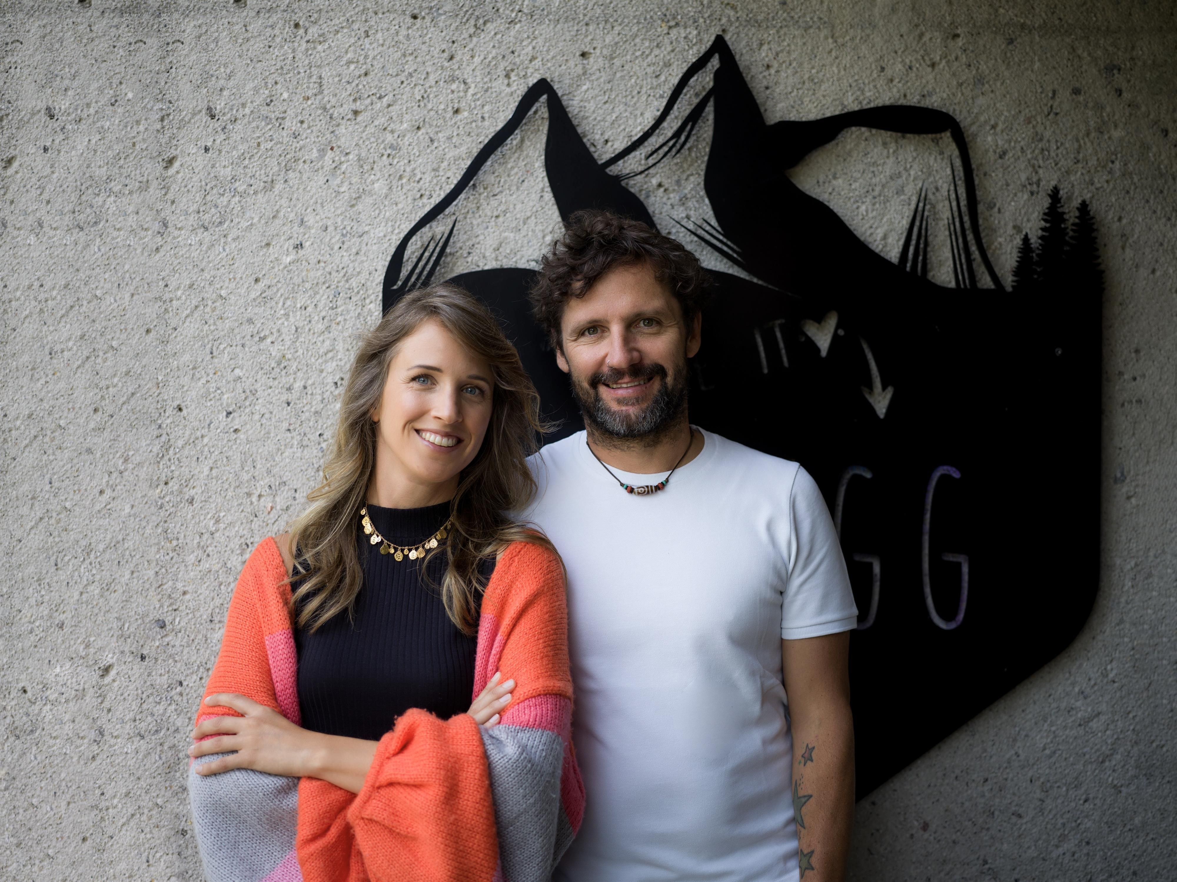 A couple is smiling in front of a gray wall with mountain motifs. The woman is wearing a colorful scarf, while the man is wearing a plain white shirt.