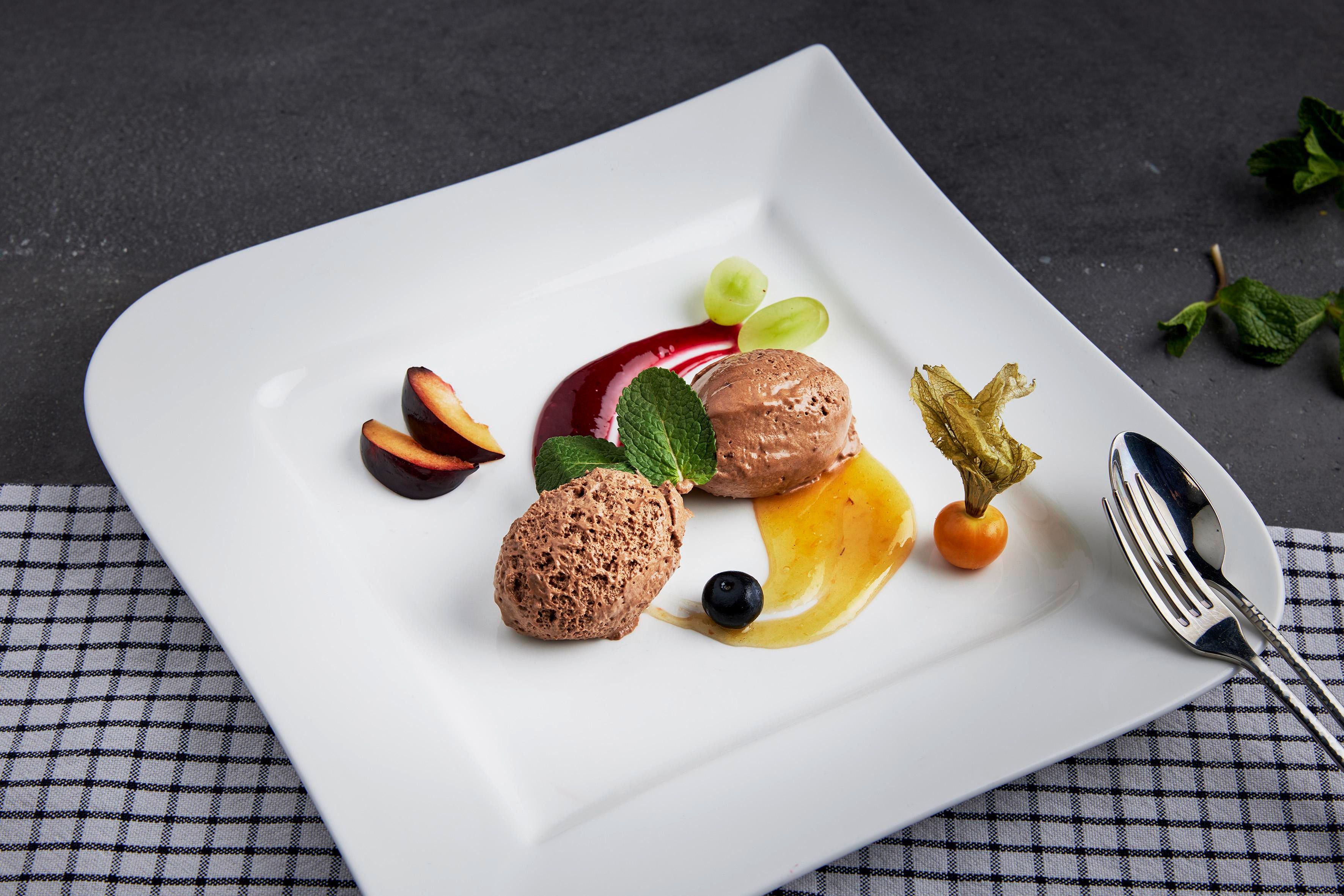 An elegant dessert on a white plate with chocolate ice cream, fruit sauce, and fresh fruits. The dish is artfully garnished with mint.