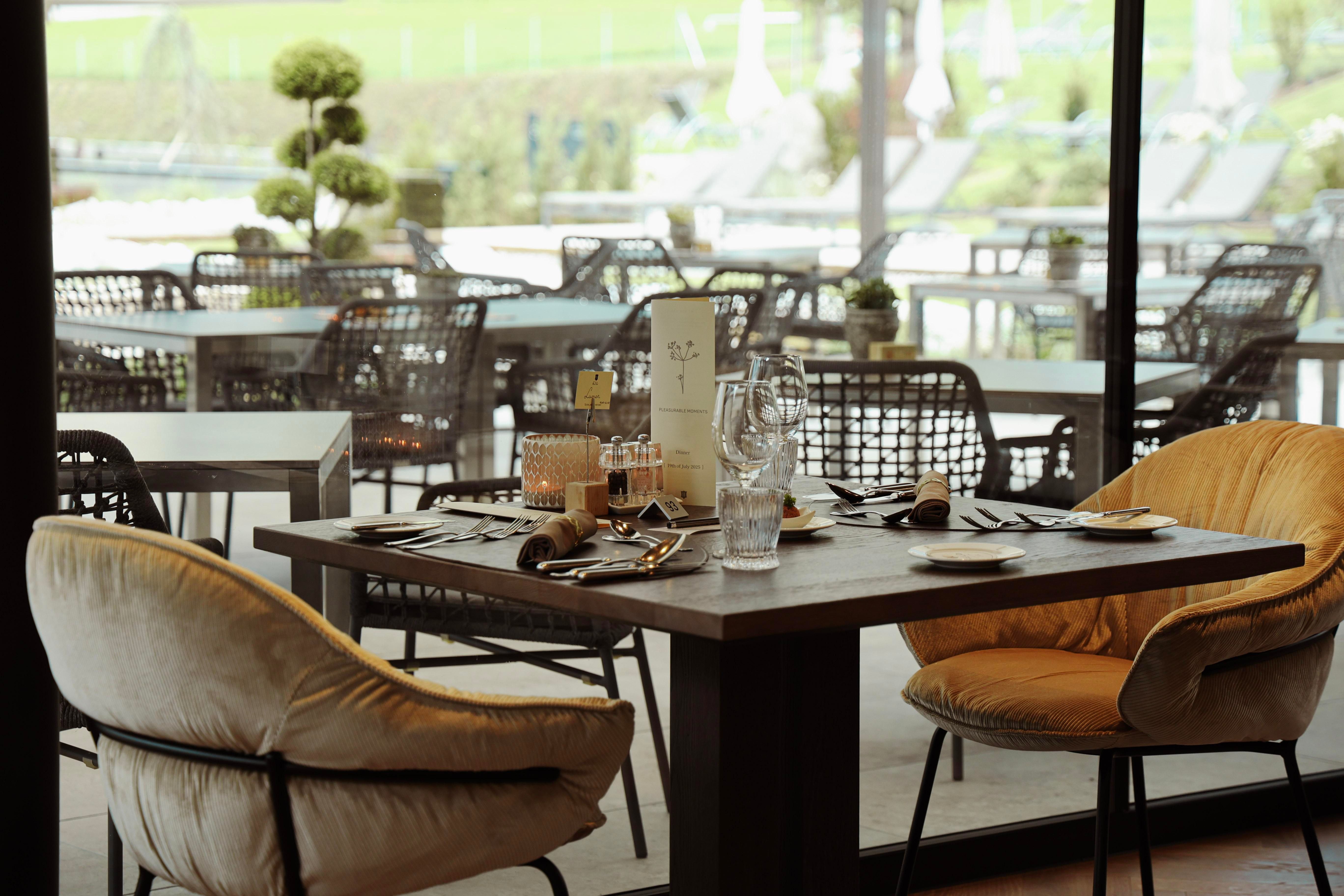 An elegant table with a cozy chair, set for a meal. In the background, additional seating and a green outdoor area can be seen.