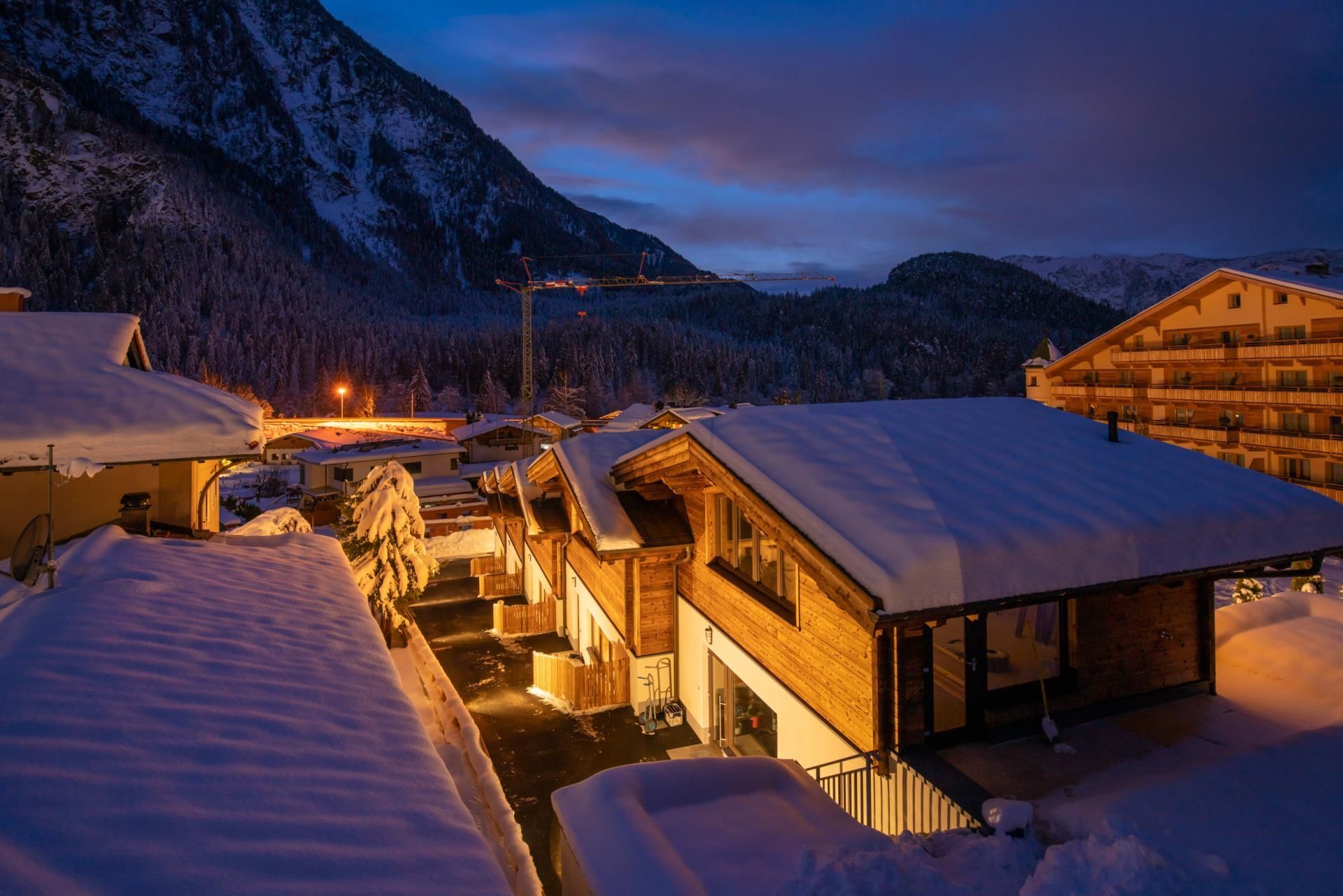 A winter landscape with snow-covered cabins and mountains in the background. The lights of the buildings shine in the twilight.